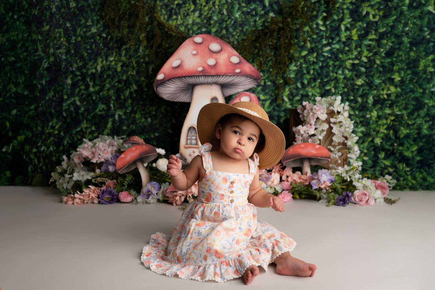A baby in a pink floral dress and wide brim hat sits among pink flowers in a garden setting.