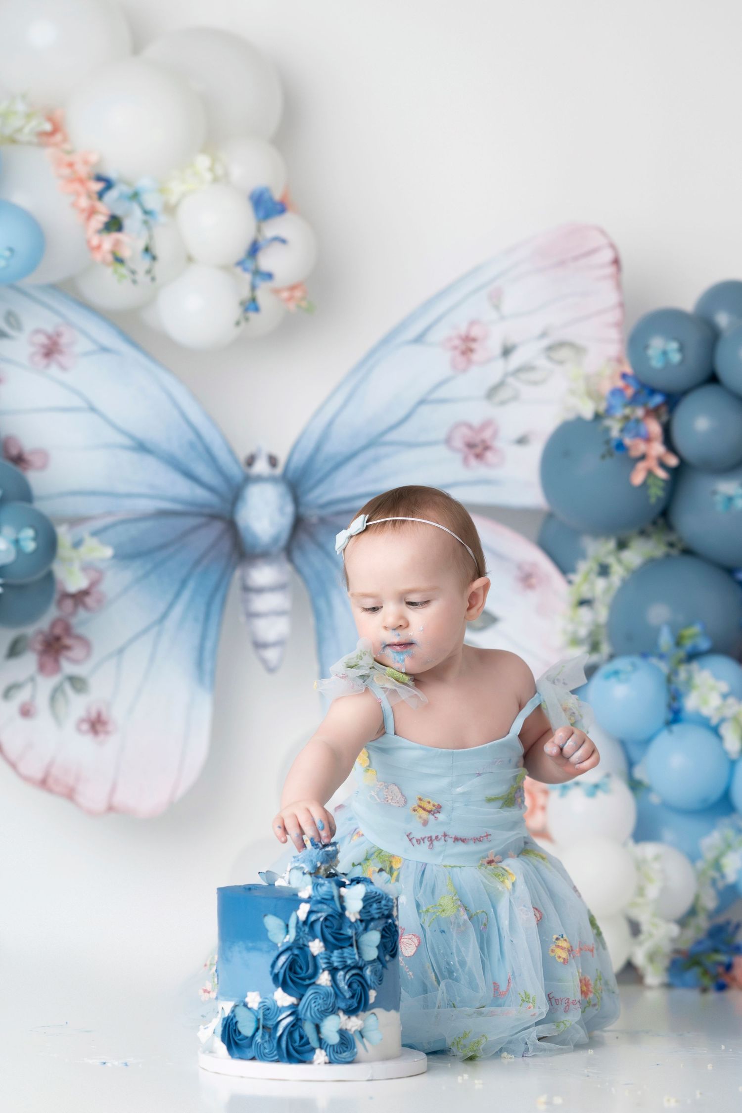 Baby in ethereal blue dress reaches for a small blue cake amidst balloon and butterfly decorations.