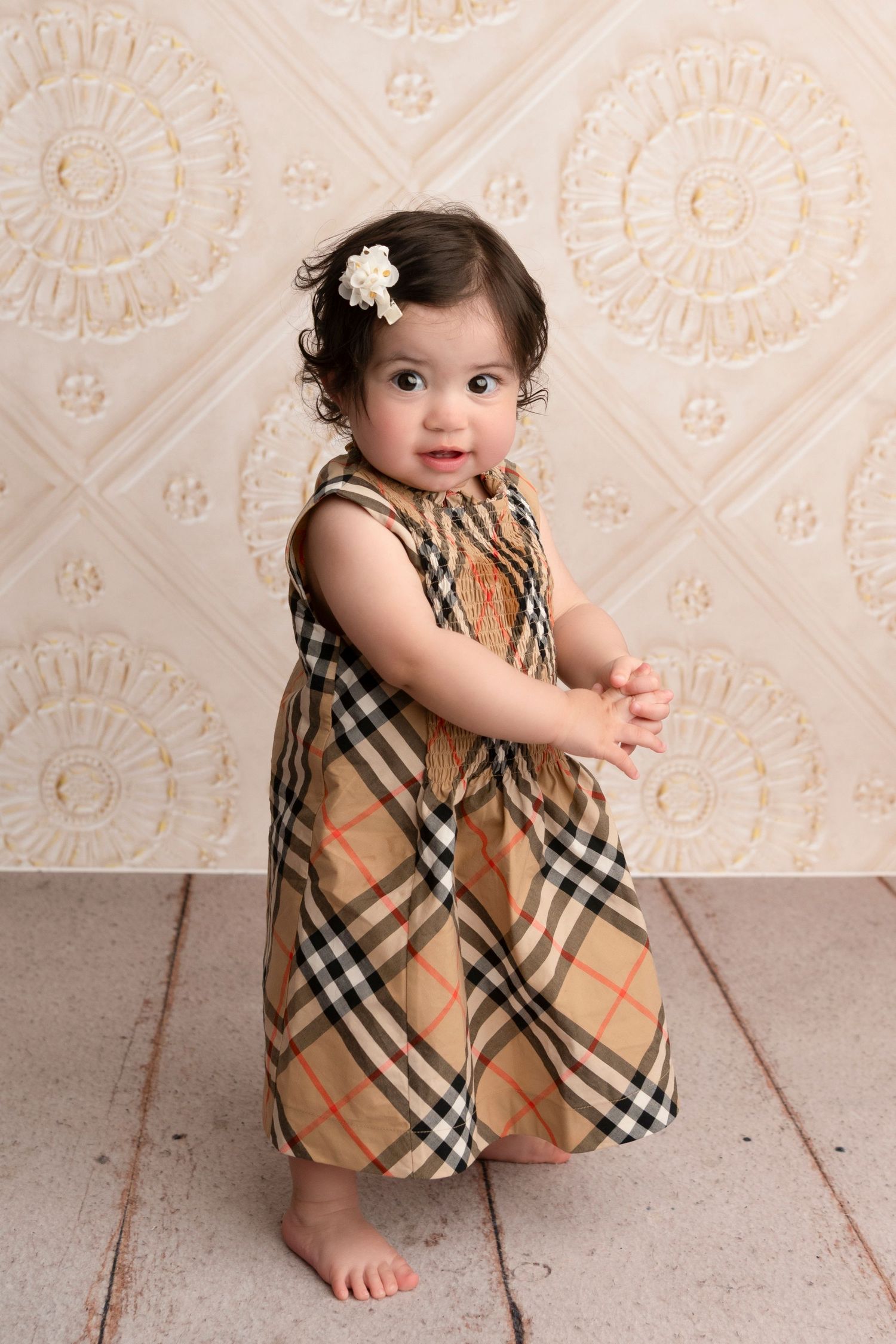 A small child wearing a plaid Burberry-style dress poses against a cream patterned wall.