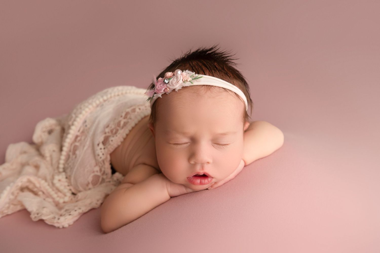 A sleeping newborn baby lies peacefully in a pink setting wearing a delicate lace headband.