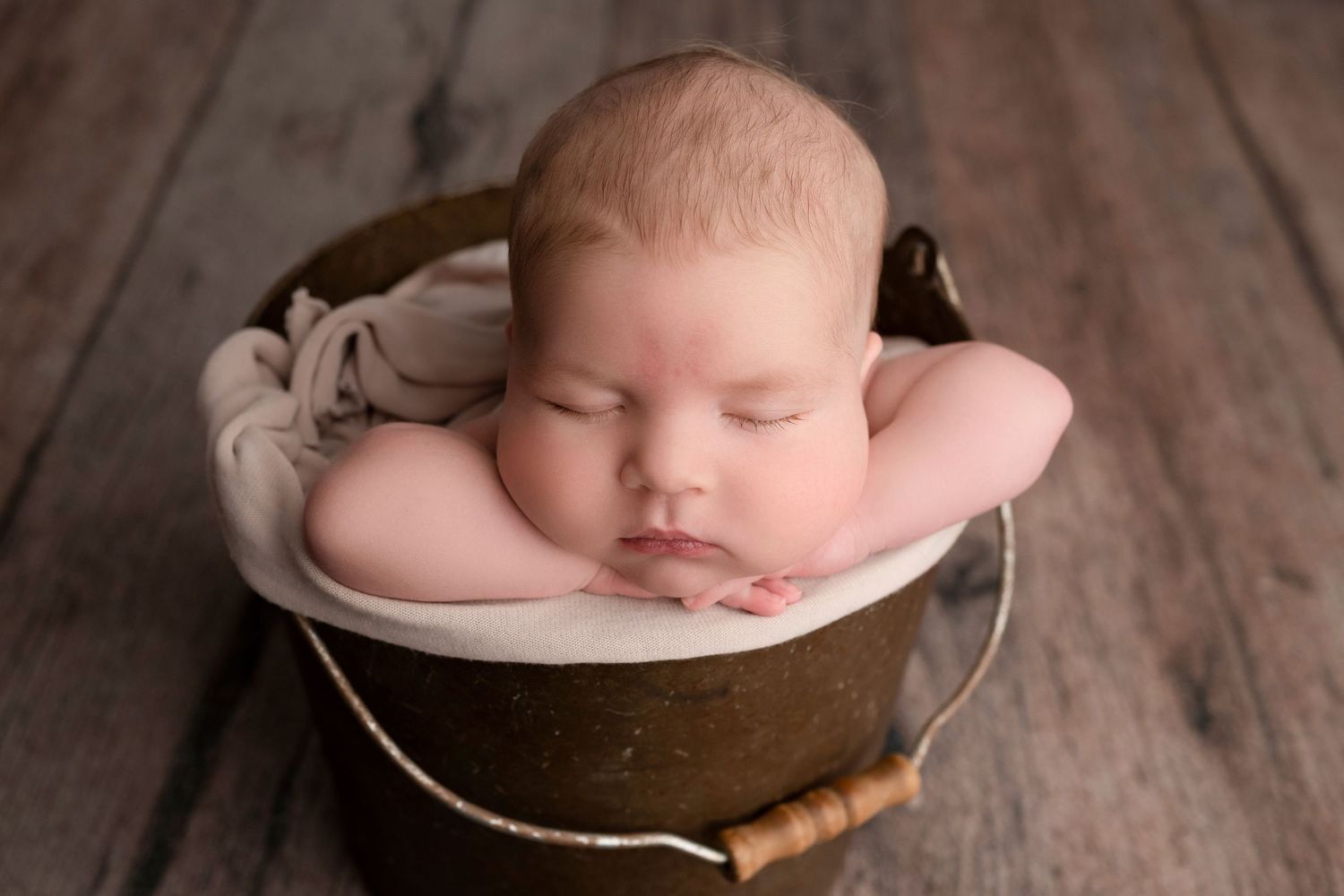 Sleeping newborn baby posed in a rustic brown bucket with cream lining on a wood floor.