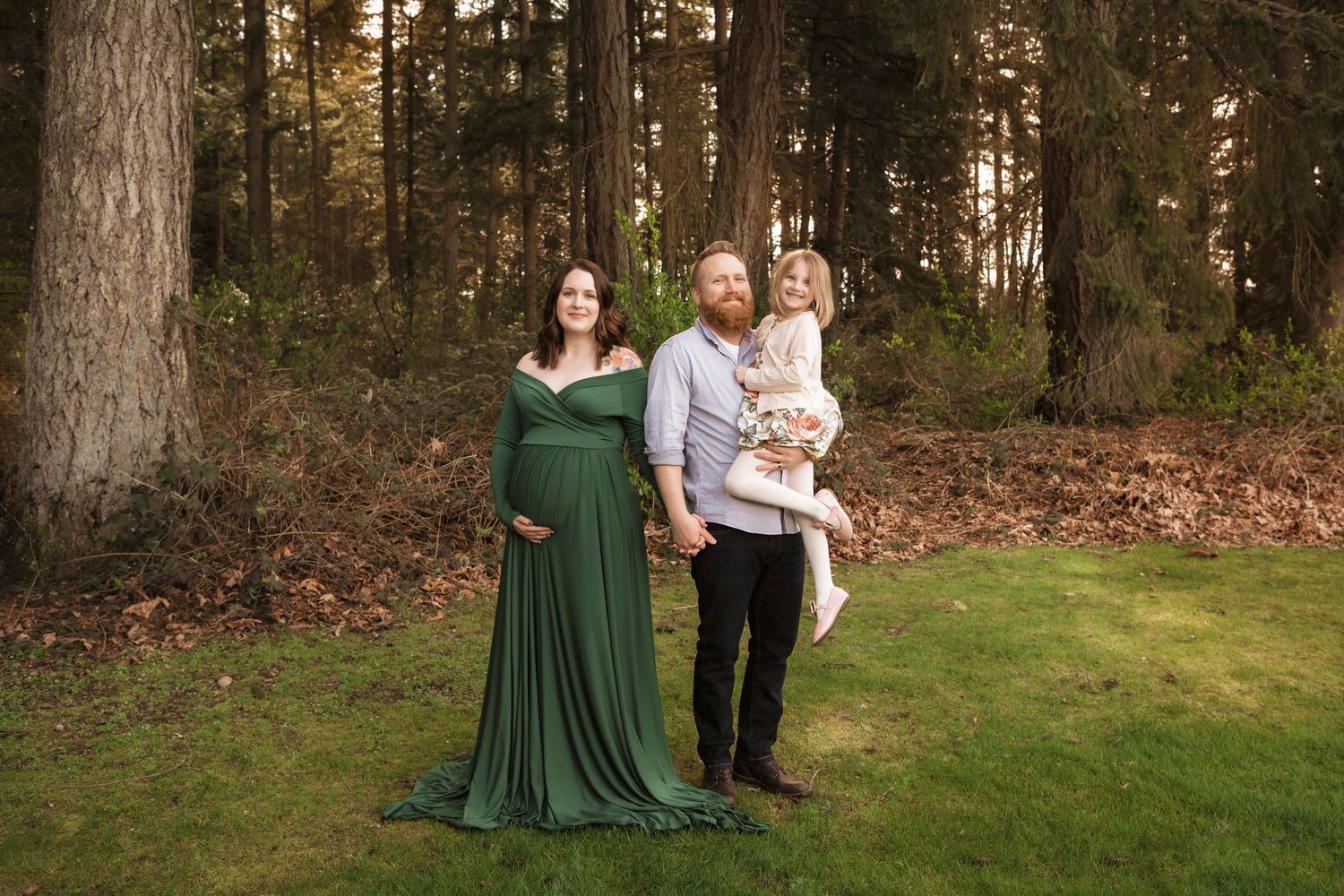 Pregnant woman in green gown with husband and toddler posing outdoors in a forest setting for maternity photos.