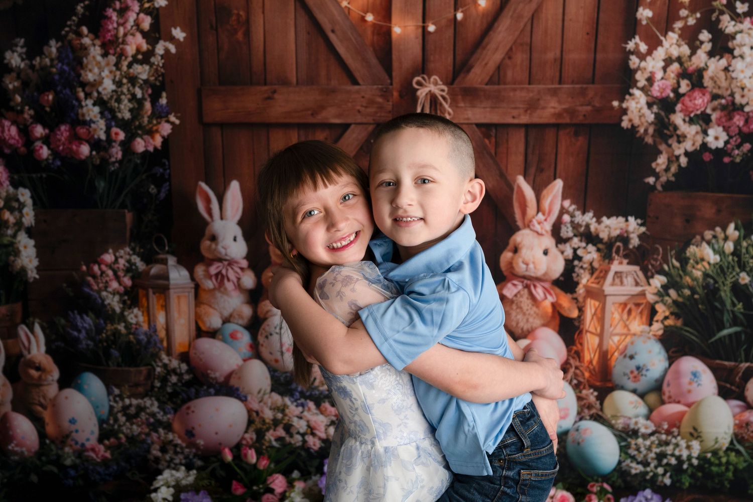 Two children hugging in front of an Easter-themed backdrop with bunnies, colorful eggs, and flowers.