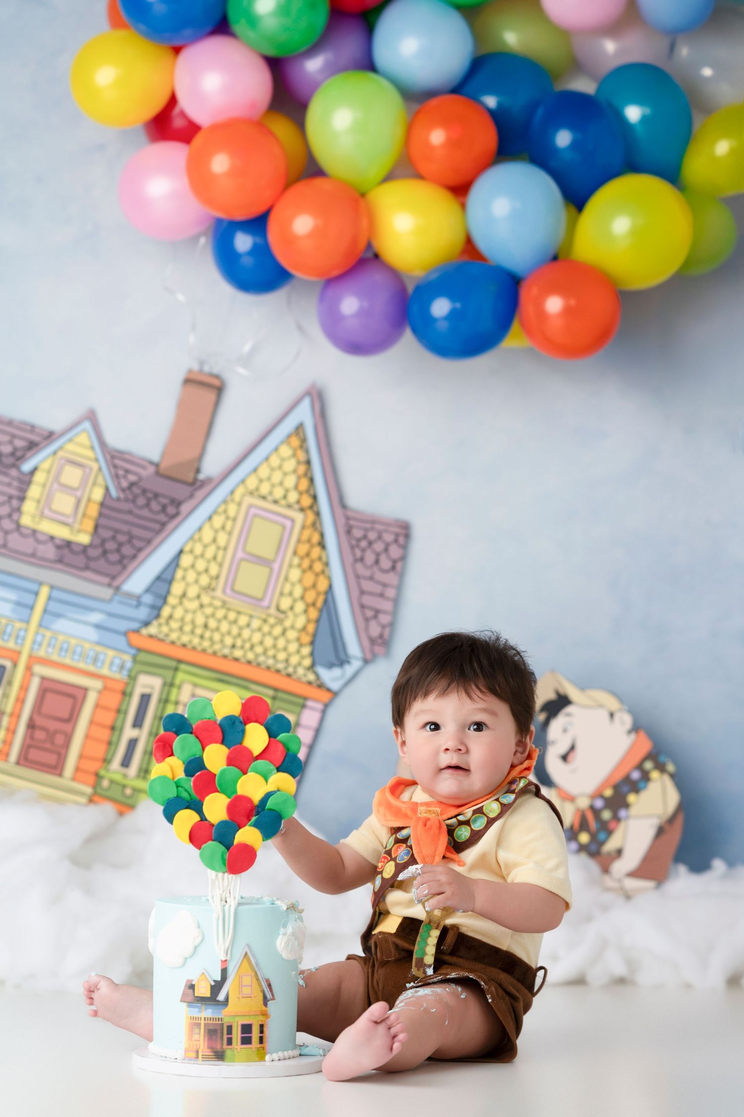 Colorful balloon garland decorates a whimsical scene with toy houses and cotton clouds in a studio setting.