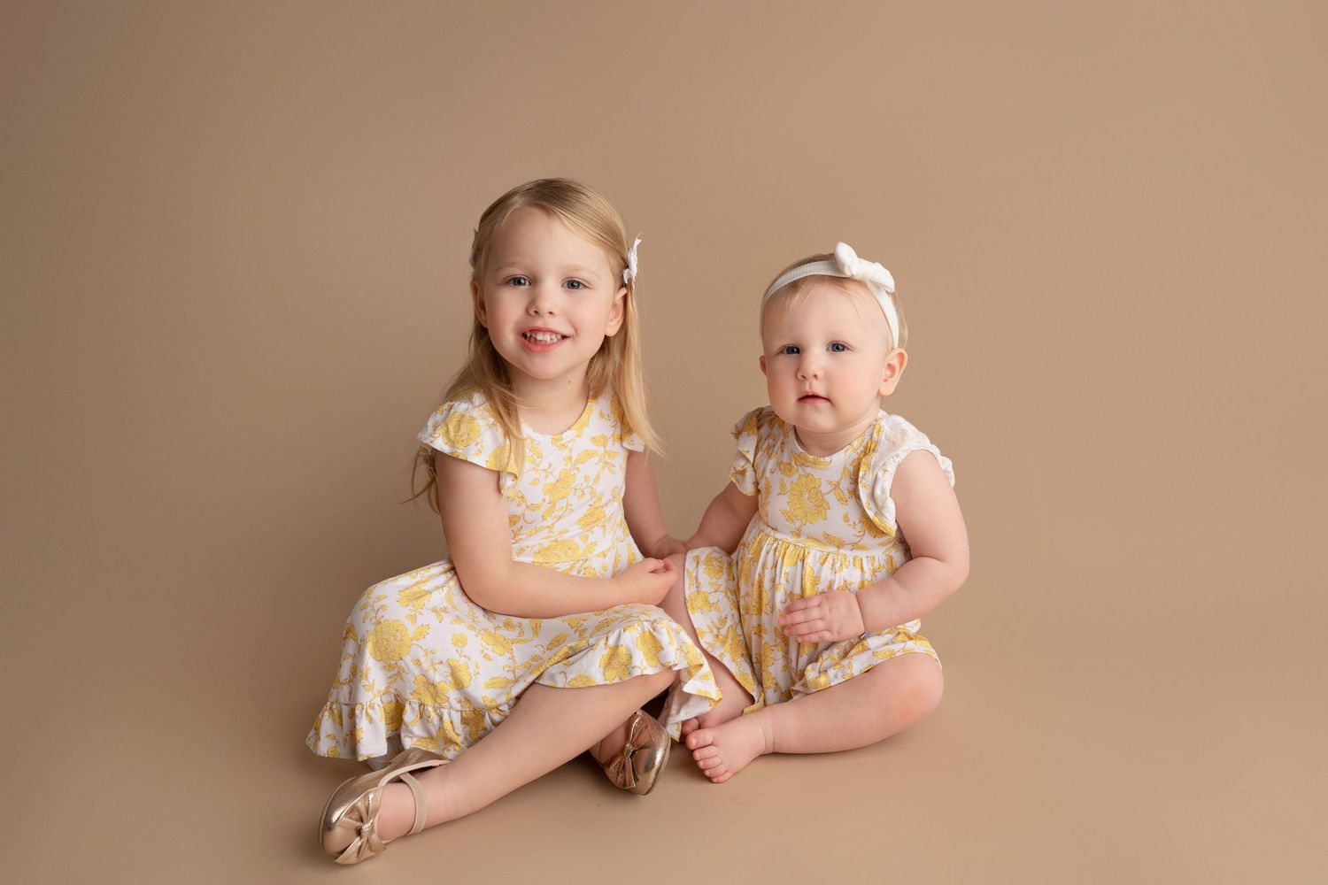 Two children wearing matching yellow floral dresses sit together on a beige backdrop for a studio portrait.