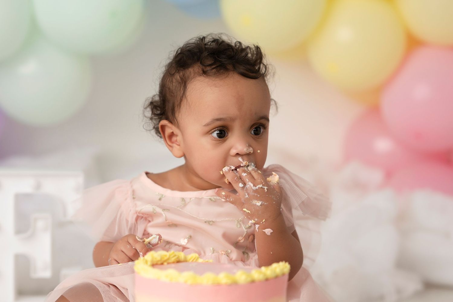 A baby in pink dress enjoying birthday cake smash session with colorful balloons in background.