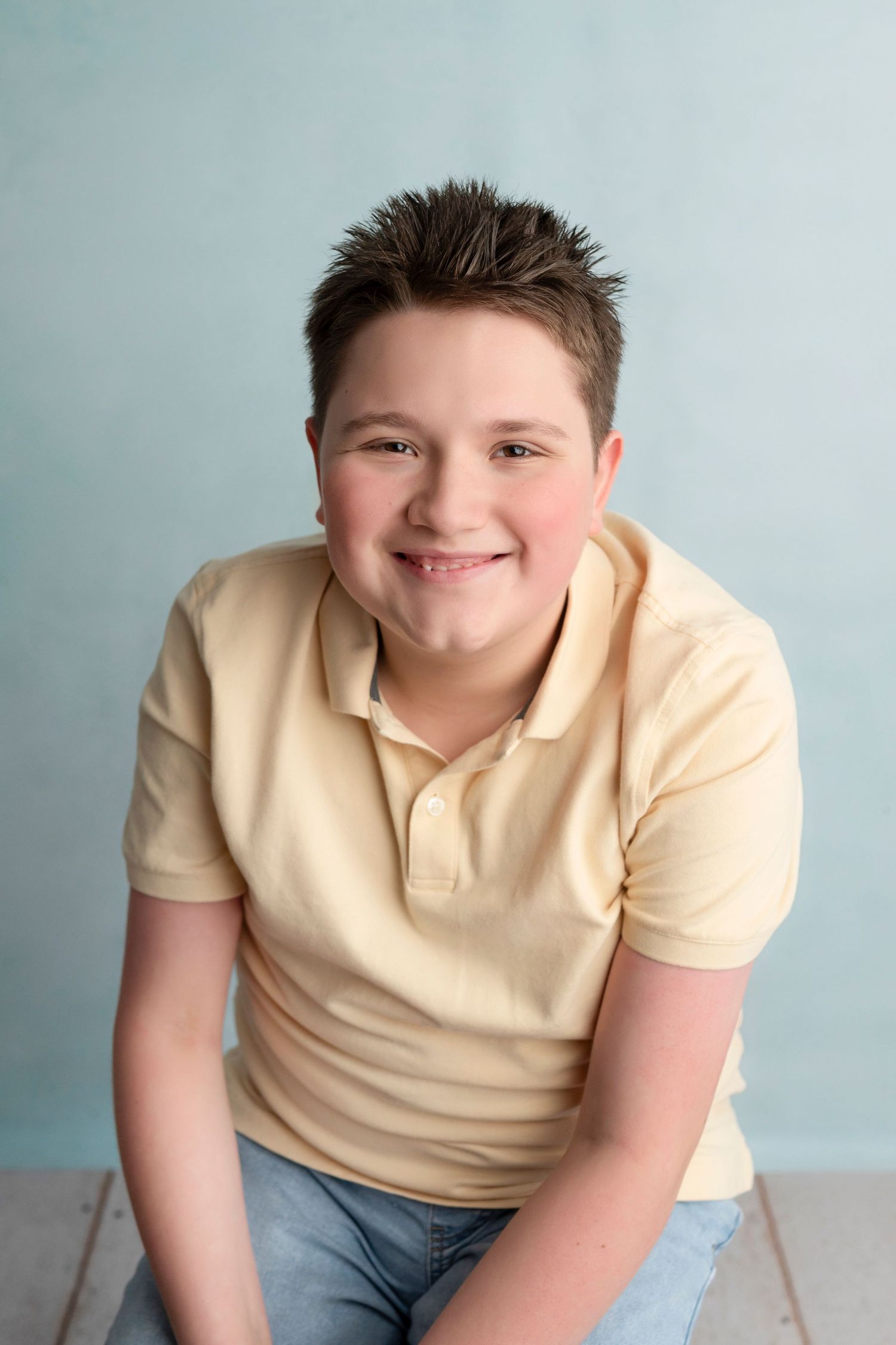 A smiling preteen boy with spiked brown hair wearing a yellow polo shirt and jeans poses for a portrait.
