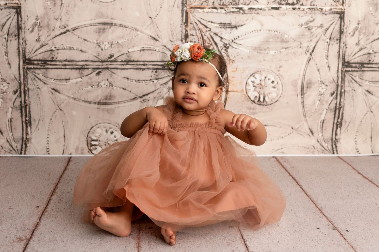 A baby girl in a dusty rose tulle dress and floral headband sits on a white wooden floor for a portrait.