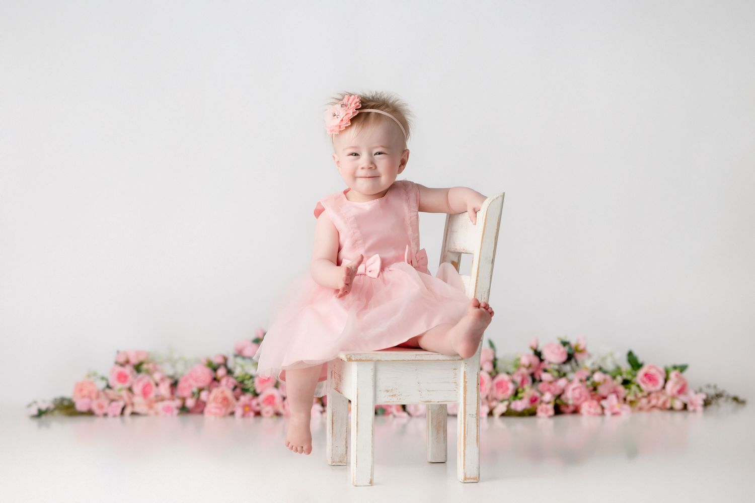 Baby girl in pink dress and floral headband smiling on white chair surrounded by pink roses.