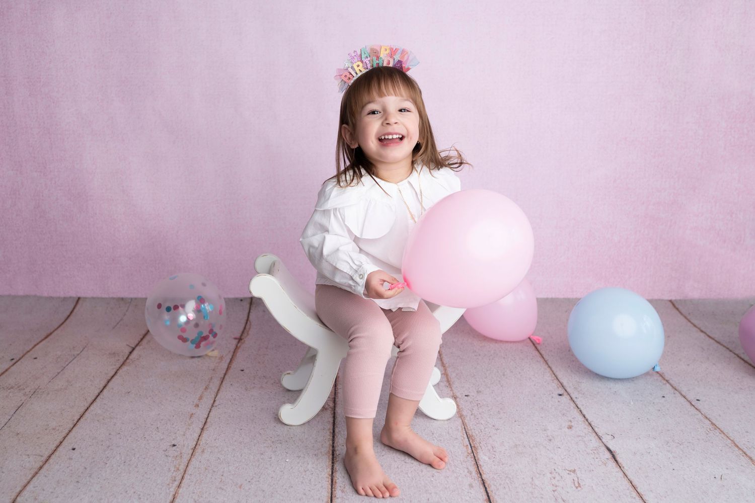 Happy young girl sitting on a white rocking horse with pink balloon, wearing flower crown on pink background.