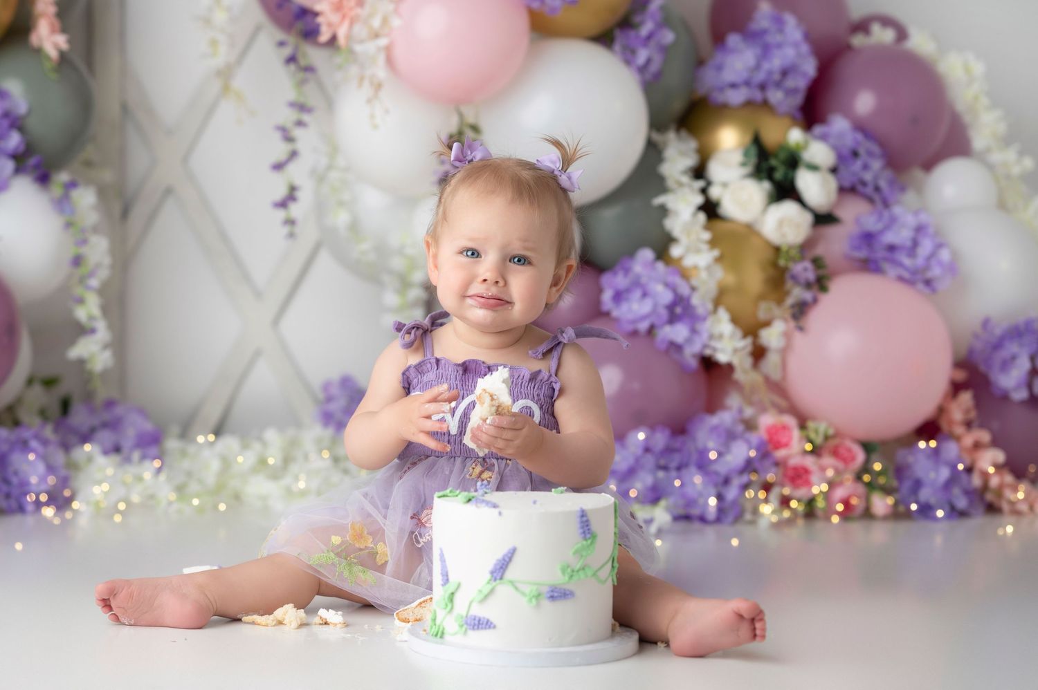 Adorable baby in lavender dress holds cookie beside first birthday cake with balloon floral backdrop.
