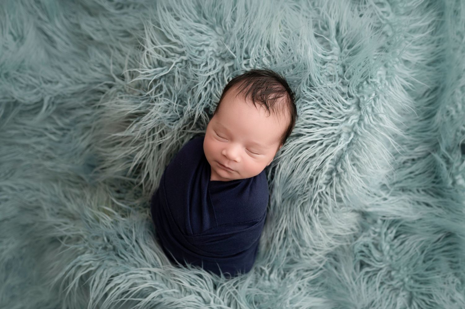 Newborn baby swaddled in navy blue wrap sleeping peacefully on a soft teal fluffy fur rug.