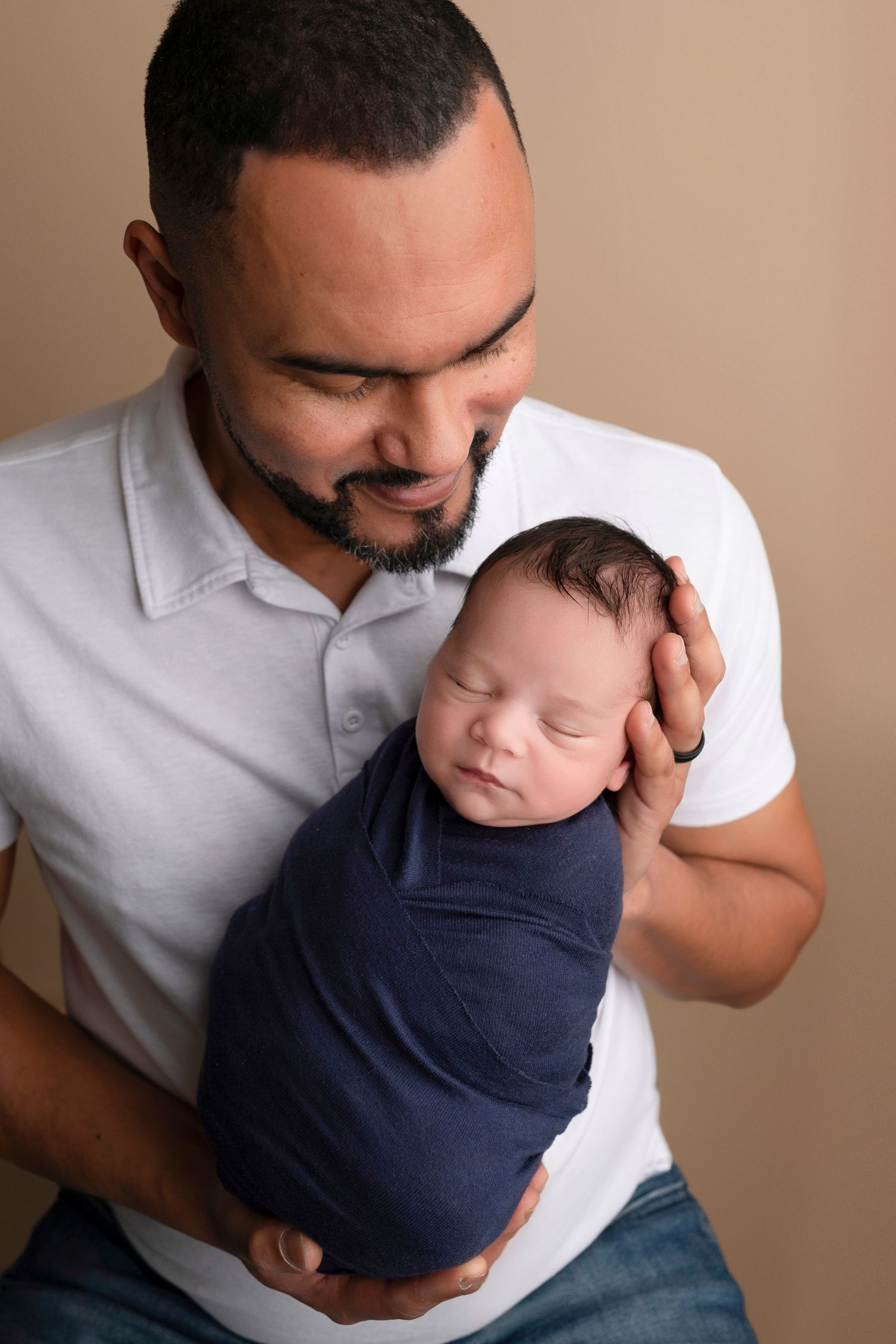 A bearded father in a white polo shirt tenderly holds his swaddled newborn baby wrapped in a navy blue blanket.