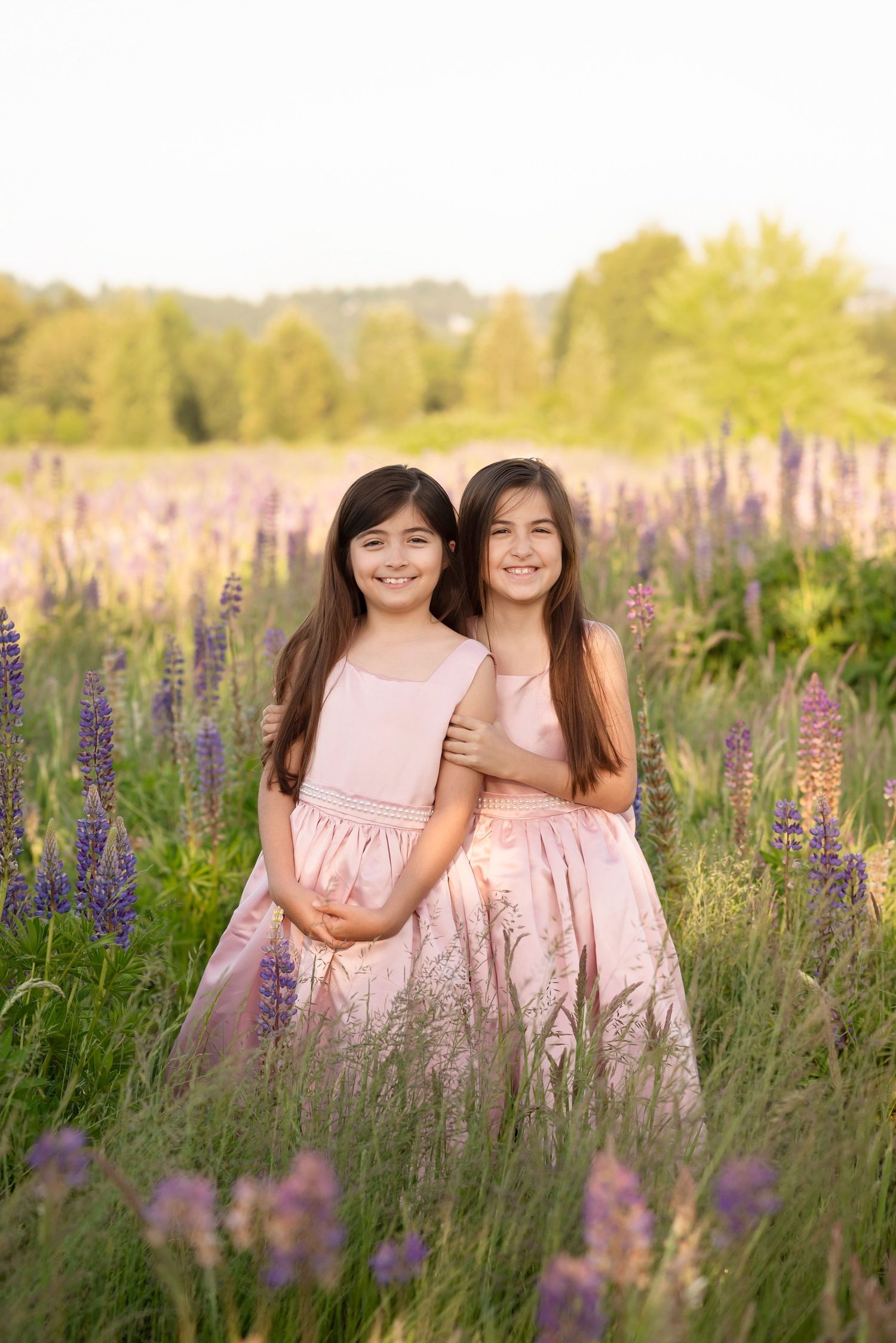 Two young girls in matching pink dresses smiling together in a field of purple lupine flowers at golden hour.
