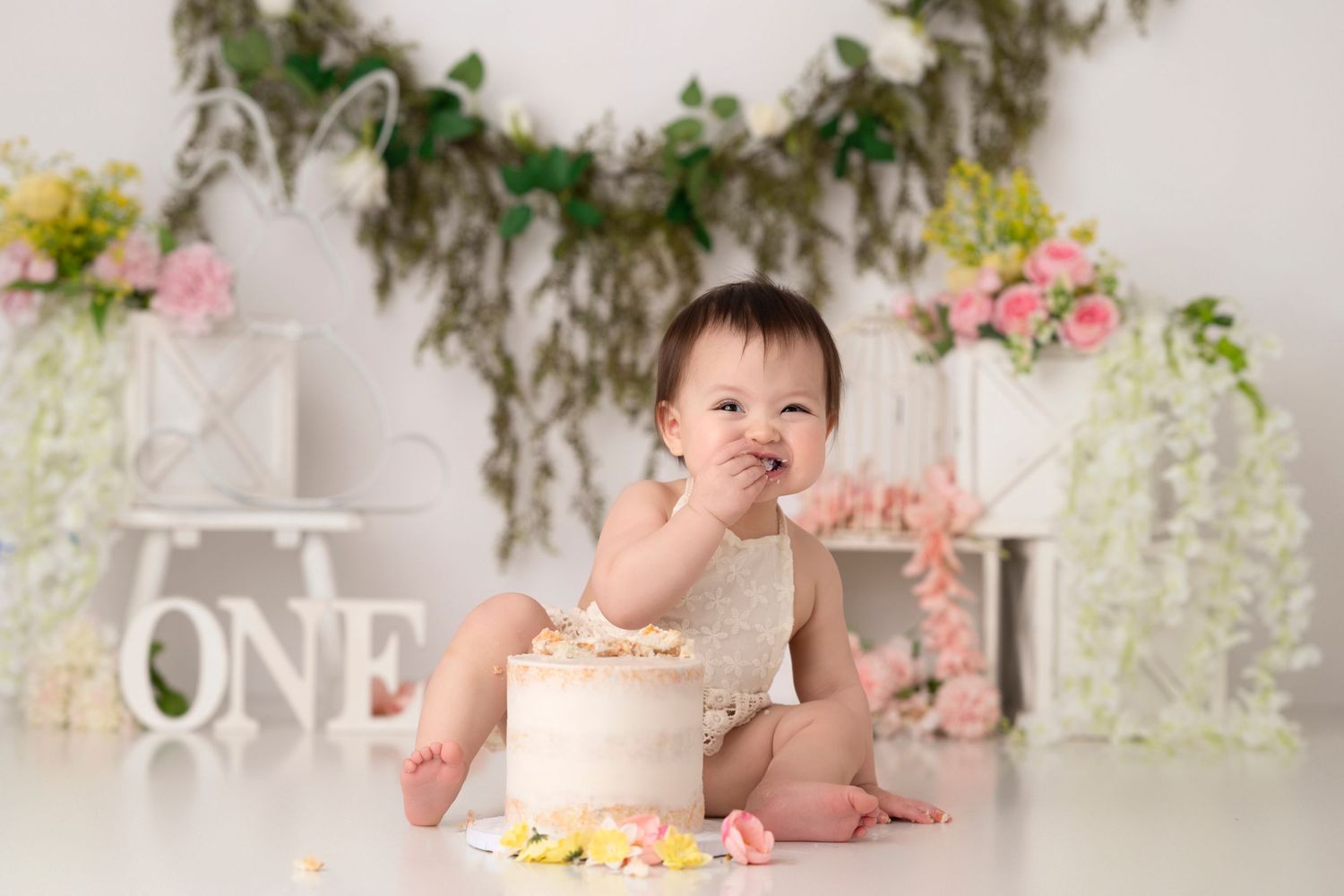 Baby sitting on floor with birthday cake during first birthday cake smash photo session with floral backdrop.