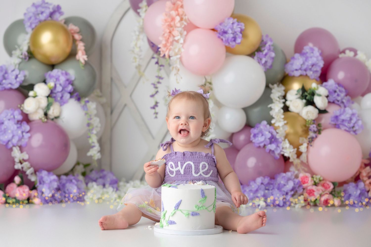 Baby girl celebrates first birthday smashing cake surrounded by purple pink gold balloons and floral backdrop.