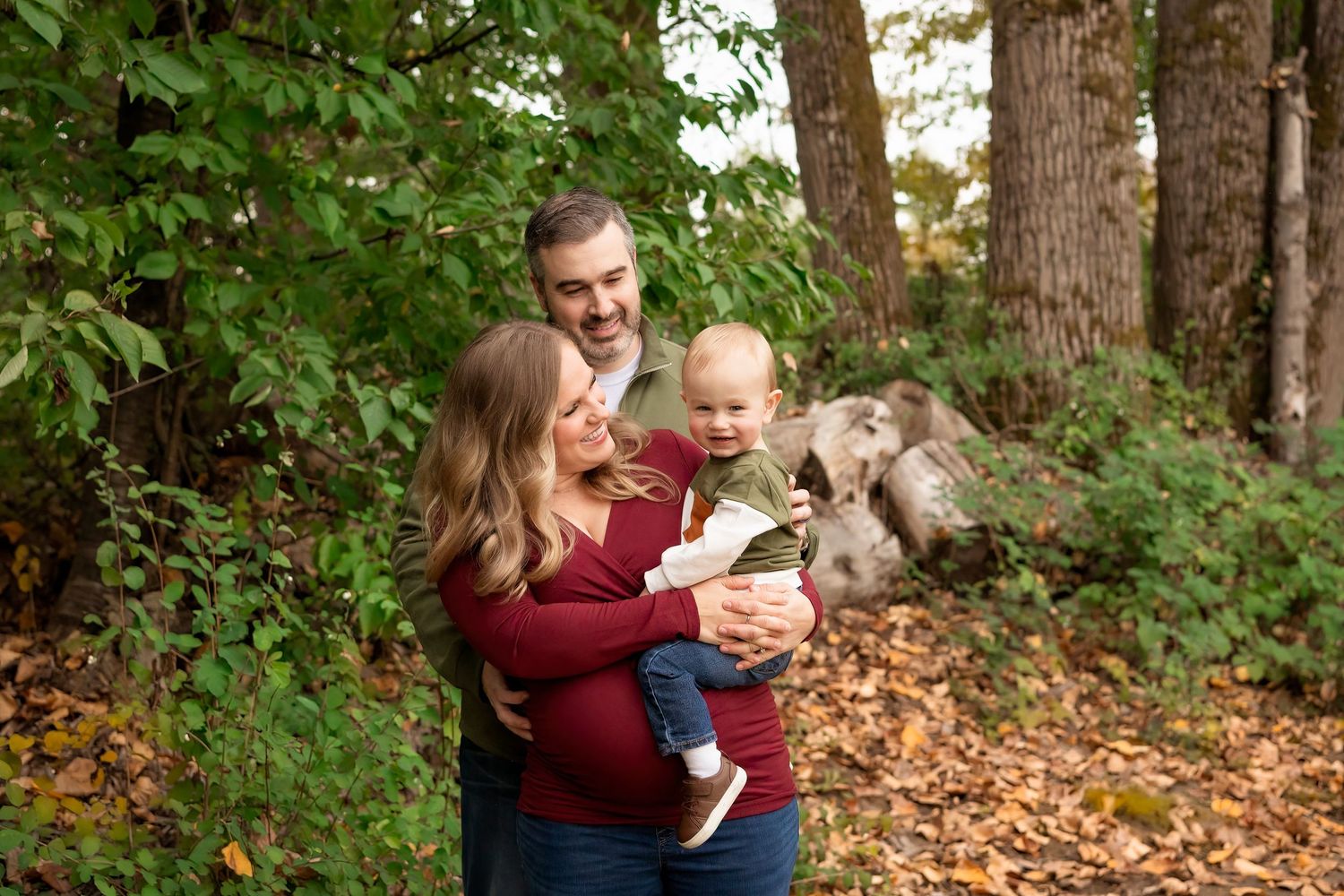 A family in maroon sweaters poses together in a wooded area with autumn leaves on the ground.