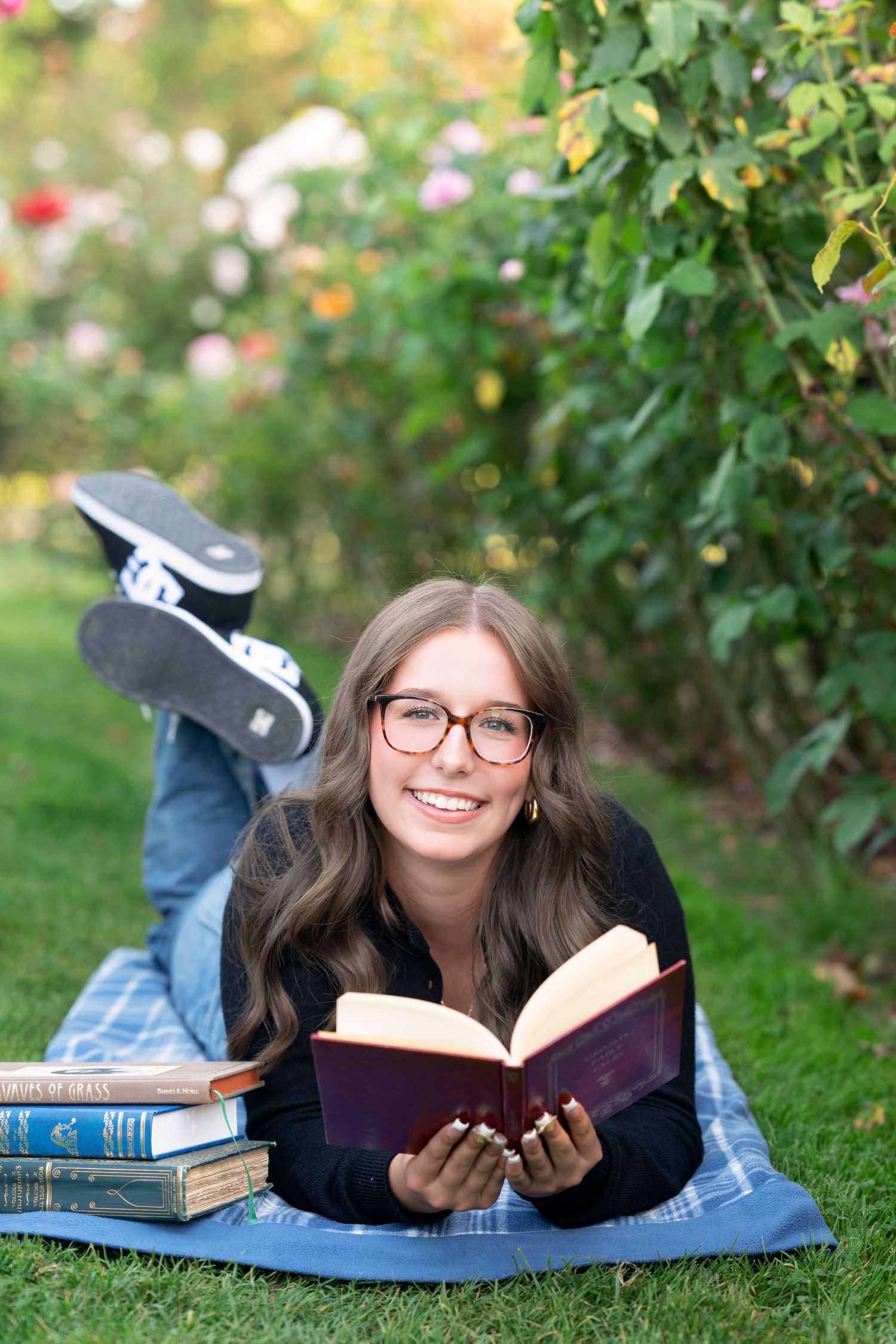 Student lying on grass reading books in a garden with blooming flowers and greenery in the background.