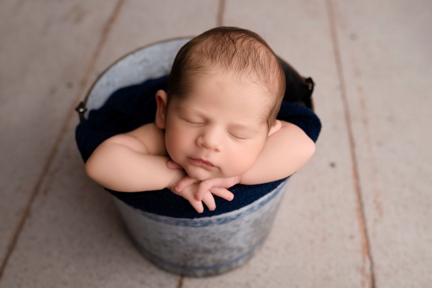 Newborn baby sleeping peacefully in grey bowl prop with navy blue wrap.