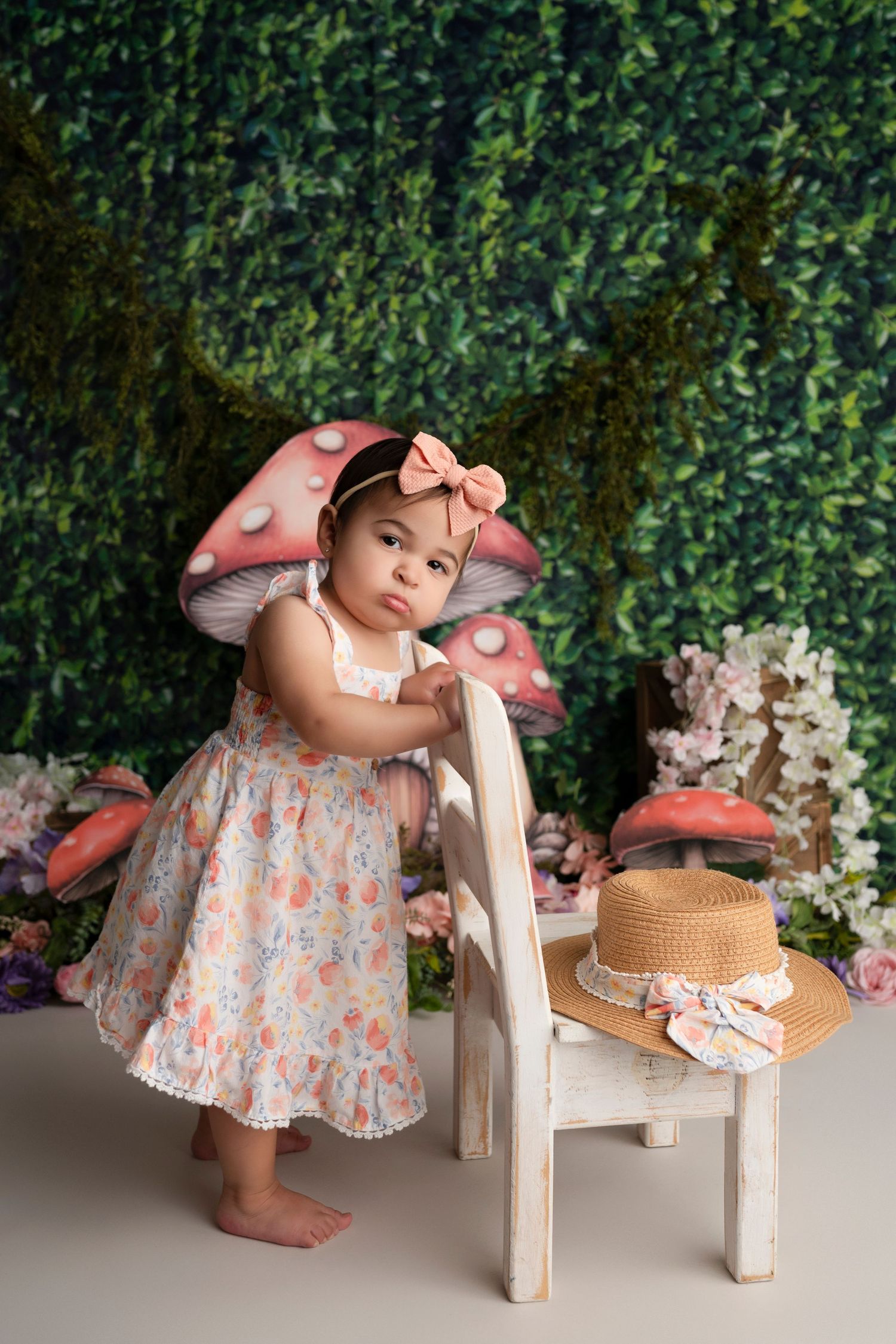Sweet photo session with small child in floral dress and straw hat against leafy backdrop with white flowers.