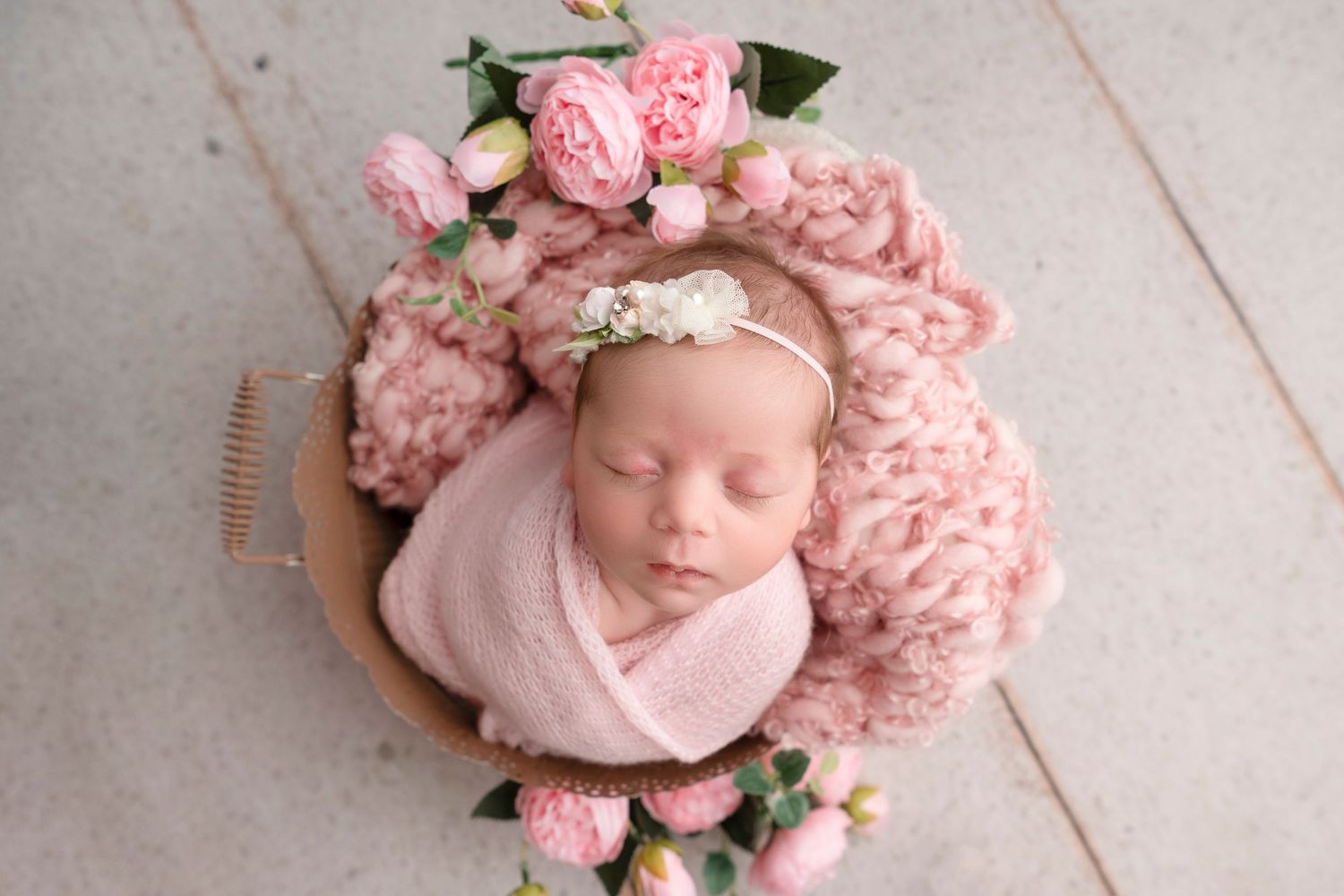 A newborn baby sleeps peacefully surrounded by soft pink flowers and delicate greenery in a professional photo setup.