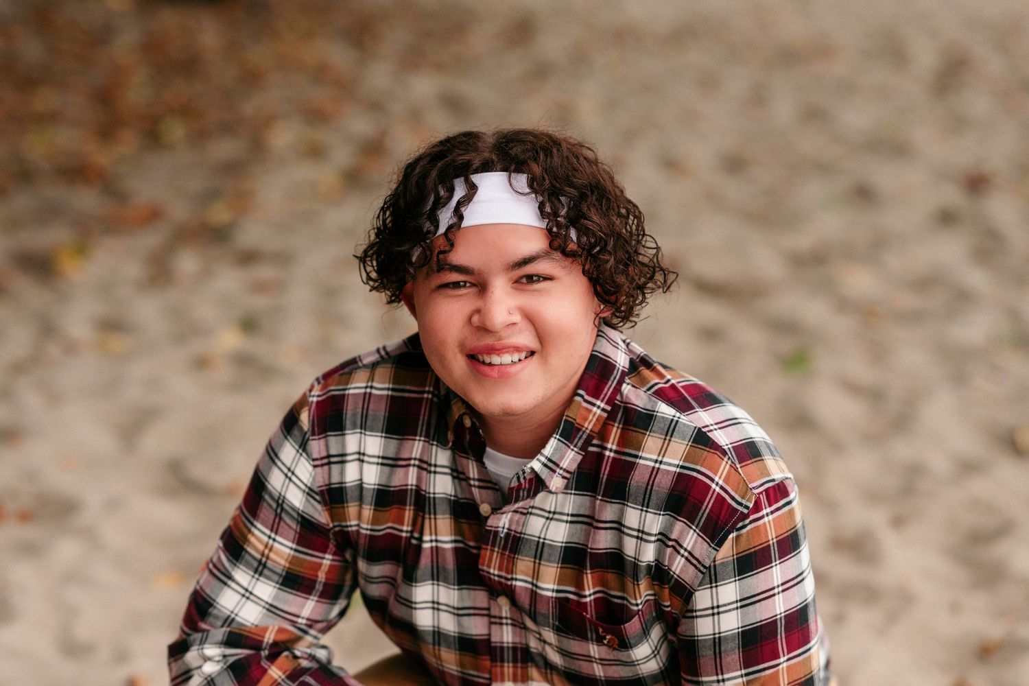 Person in plaid shirt and khaki pants poses outdoors with curly hair and headband.