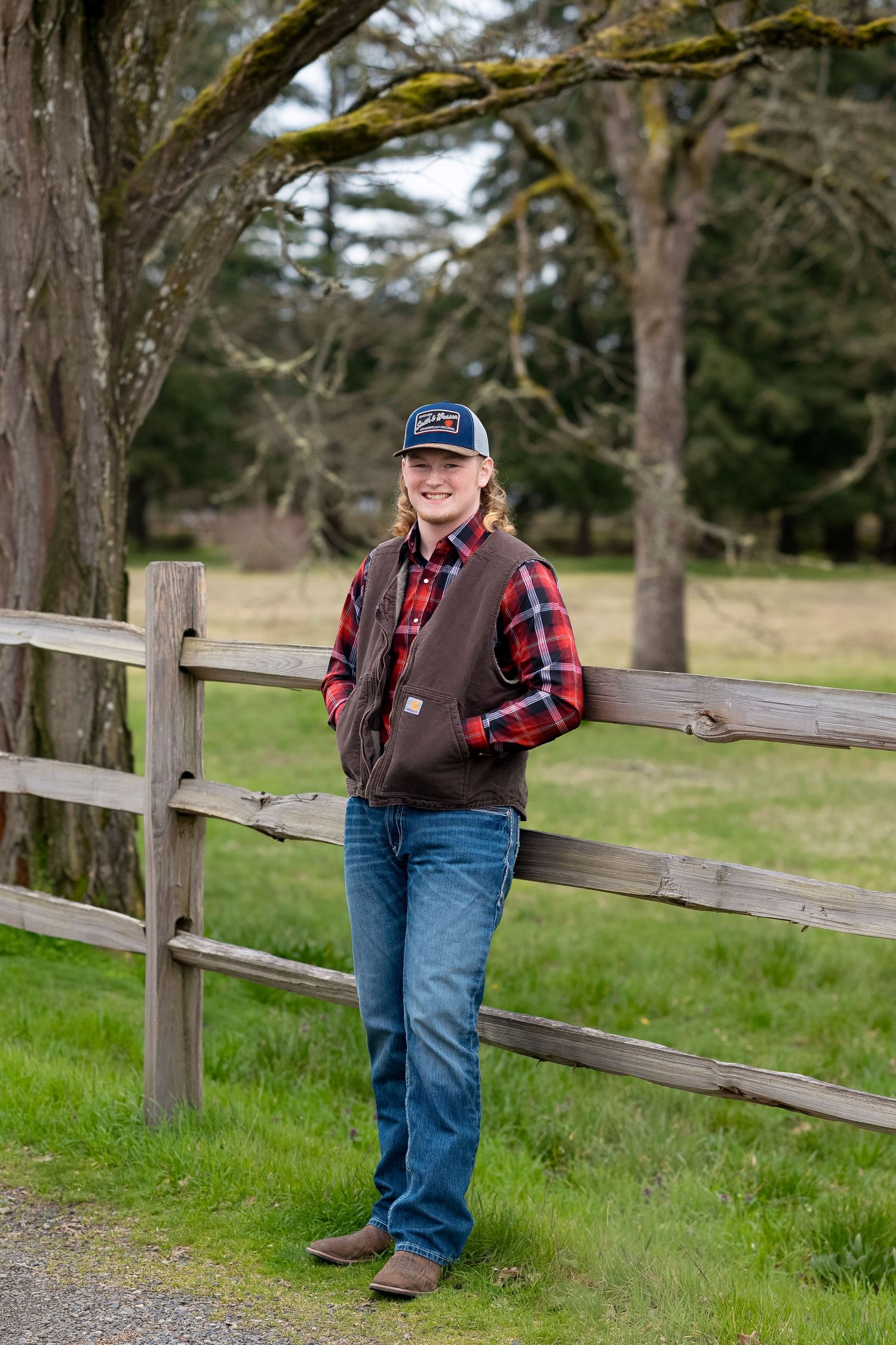 Person in plaid shirt and vest leans against rustic wooden fence in rural countryside setting.