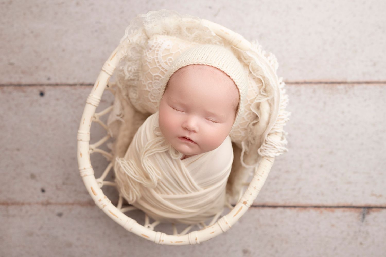 Newborn baby wrapped in cream fabric sleeping in a wicker basket wearing a knit bonnet.