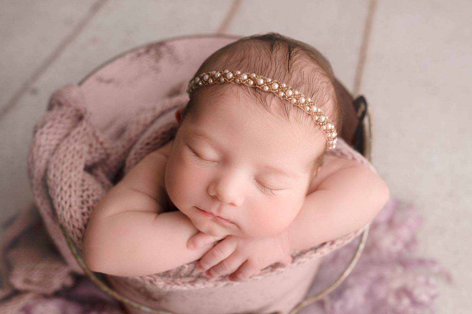 Sleeping newborn baby girl in a metal bowl wearing a gold pearl headband, wrapped in a soft pink knit blanket.