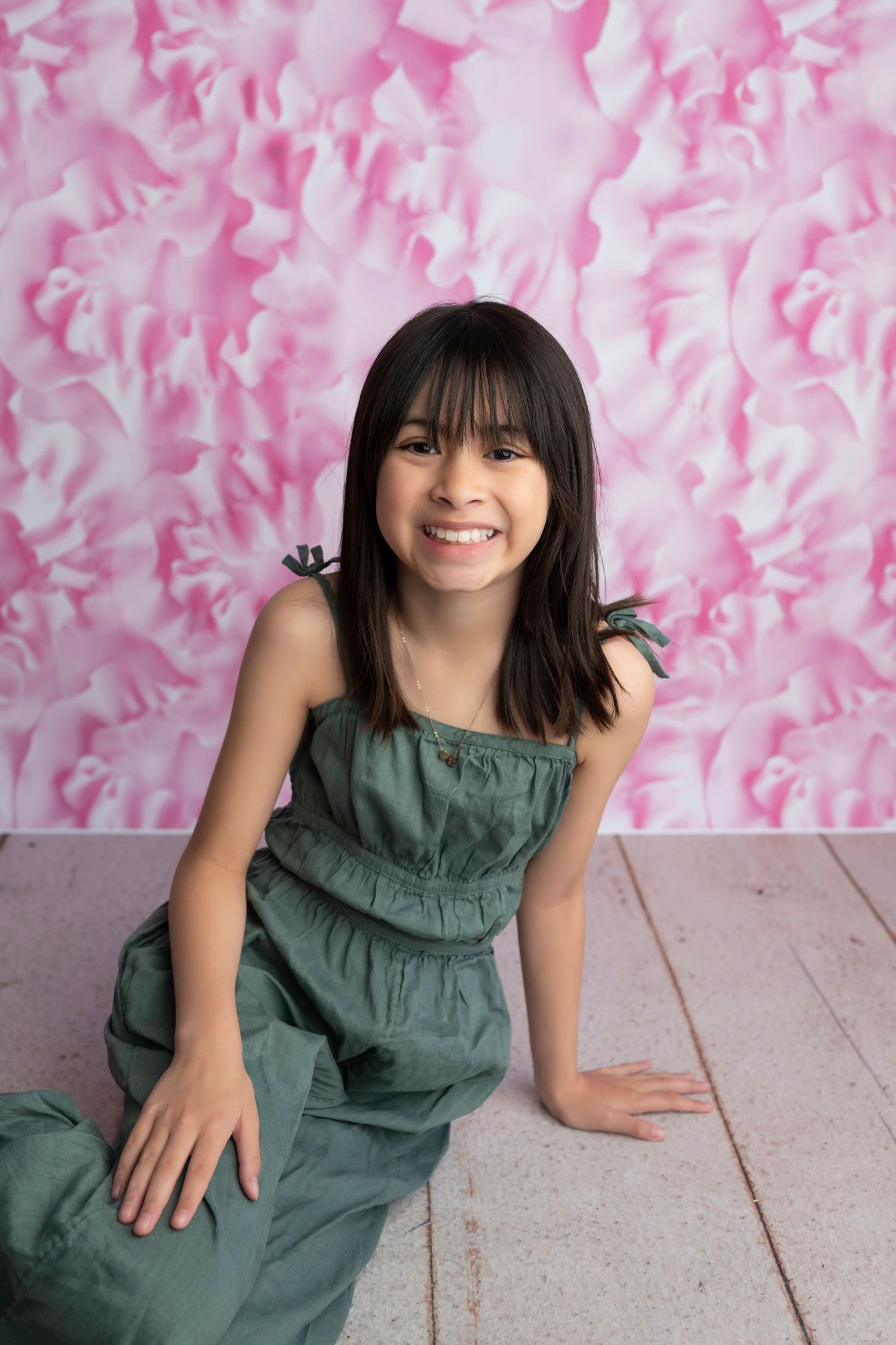 A smiling young girl in a green dress sits on a white floor against a pink floral backdrop.