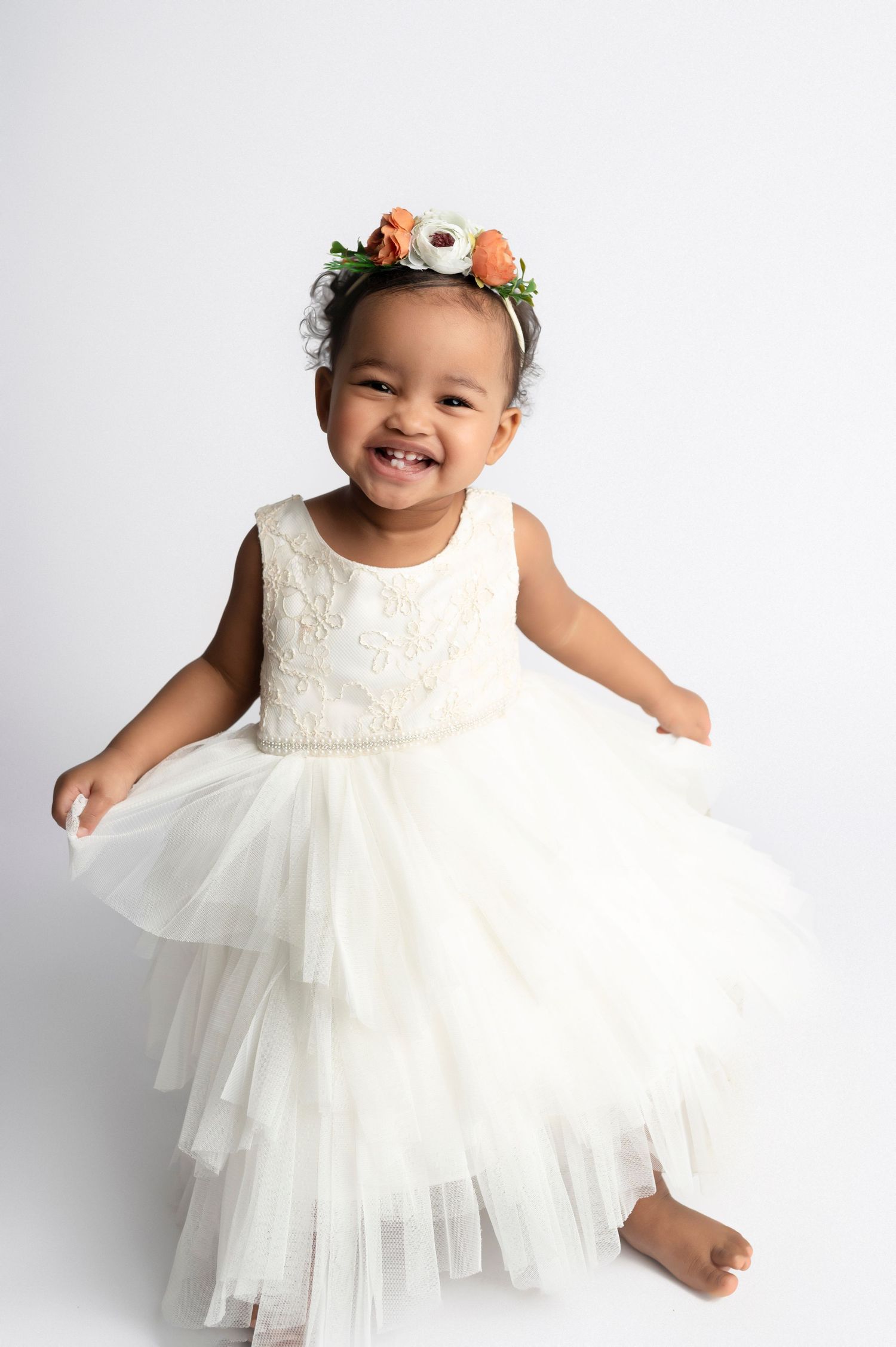 Smiling toddler girl in white tiered tulle dress and floral crown poses against white background.
