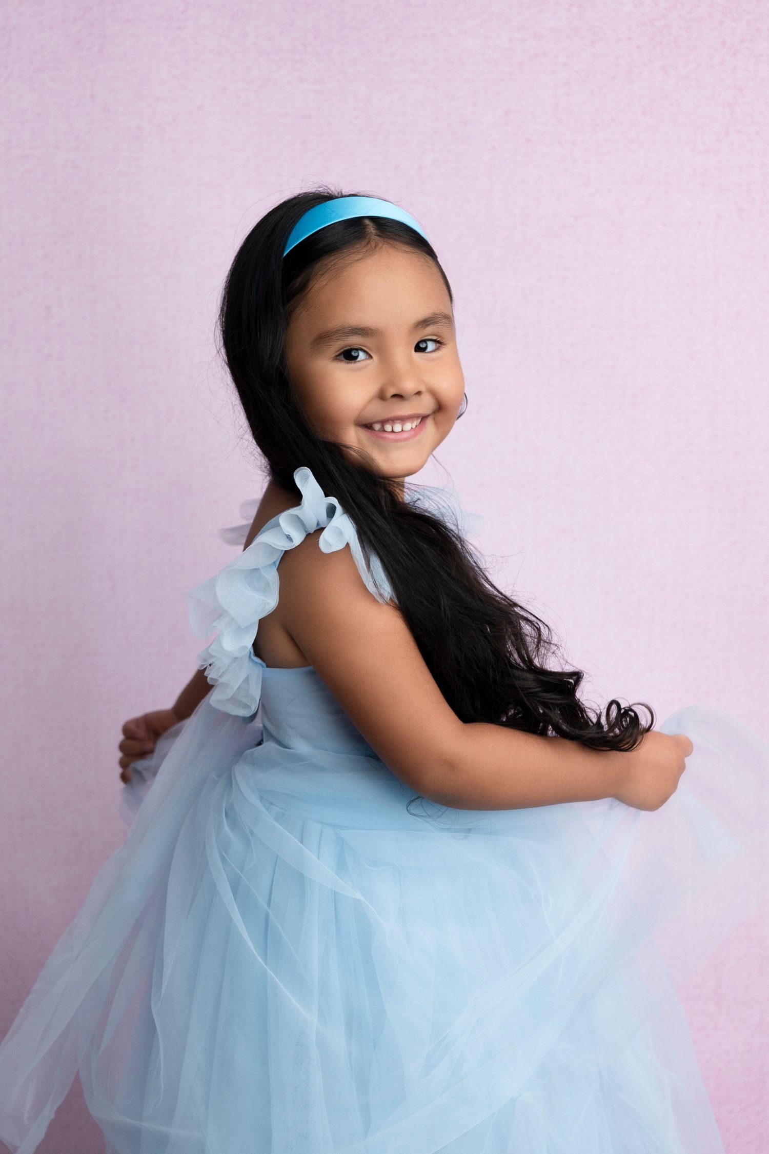 A smiling young girl in a light blue tulle dress and matching headband poses gracefully against a pink background.