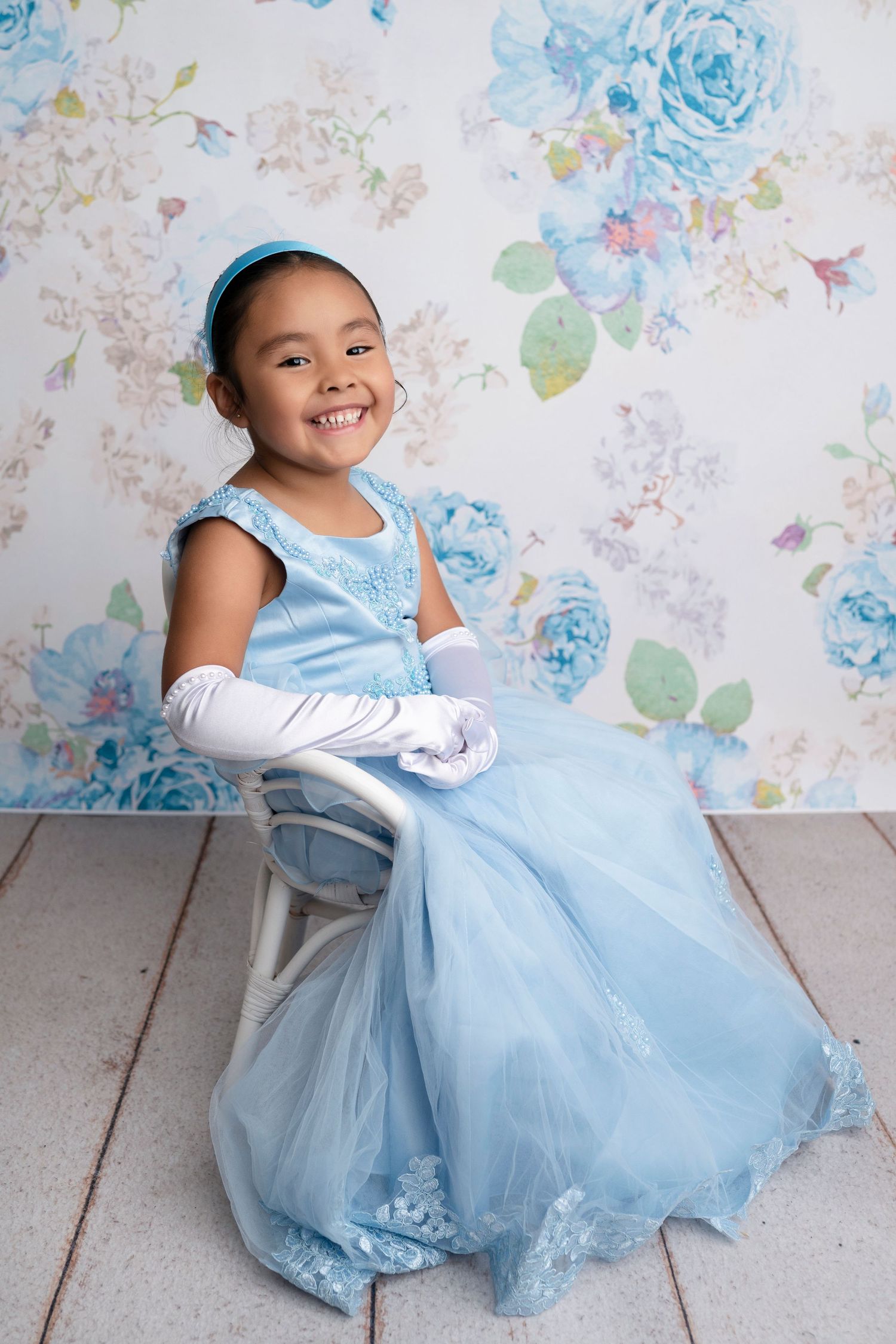 A smiling young girl in a blue princess gown and white gloves sits on a chair before a floral backdrop.