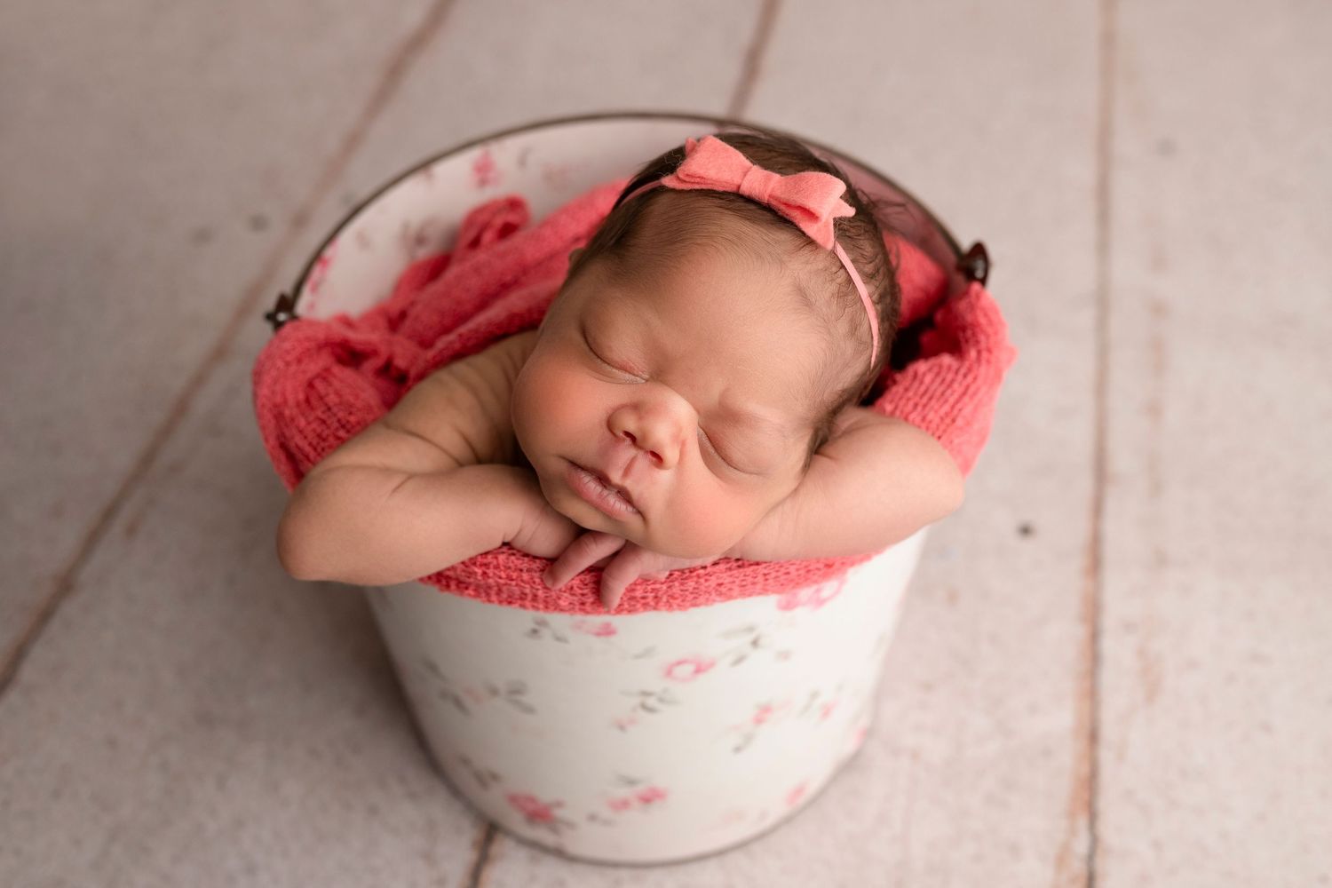 Newborn baby sleeping peacefully in a white floral bucket with pink wrap and bow headband.