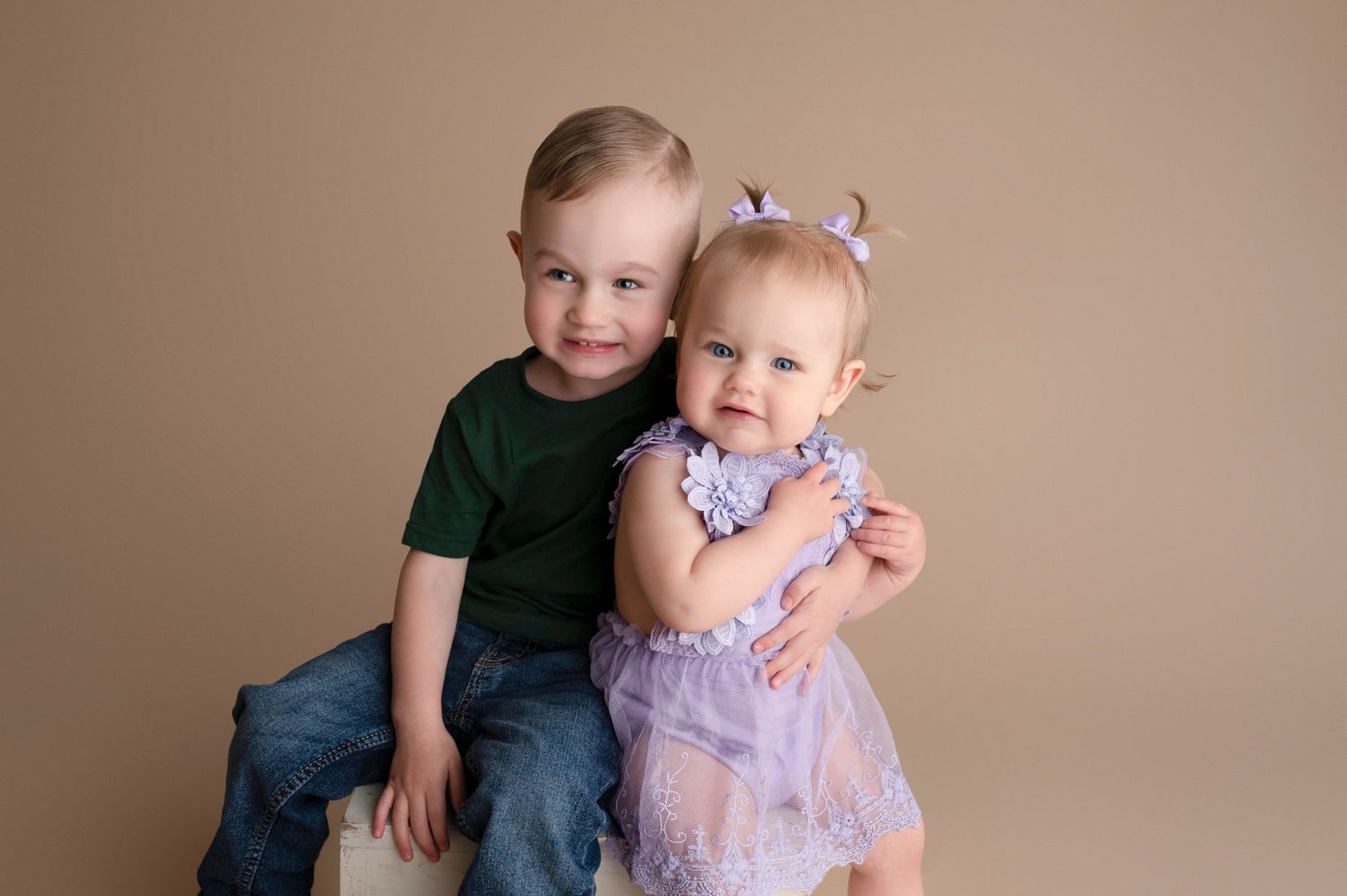 A smiling toddler boy in green shirt hugs a baby girl in purple floral dress against a beige backdrop.