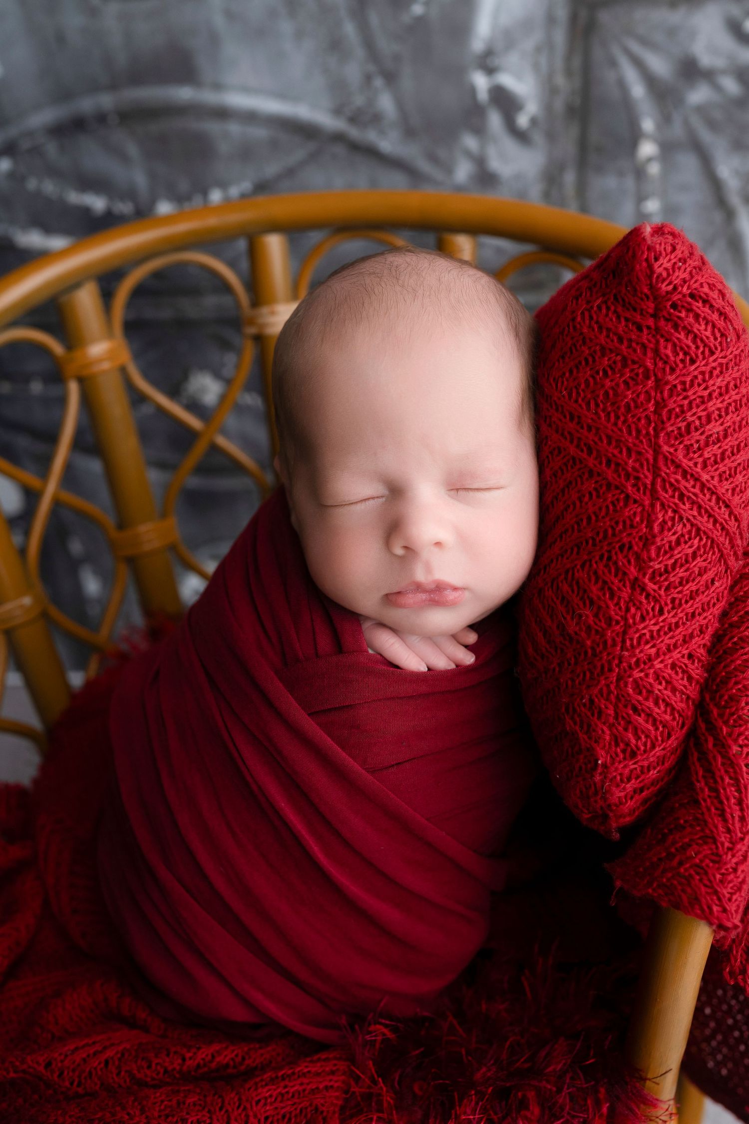 Sleeping newborn baby swaddled in red wrap sitting in a wicker chair with red knit blanket.