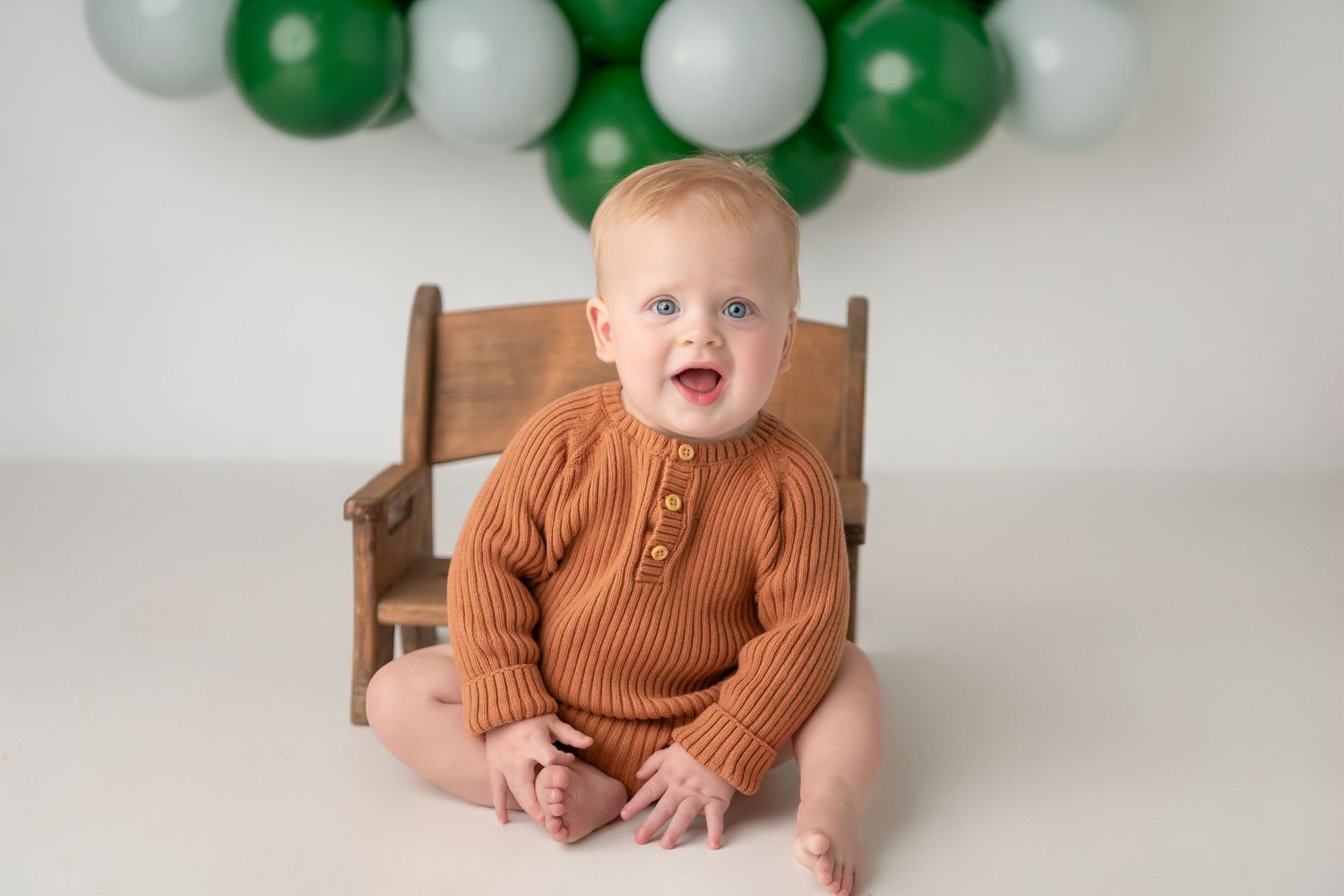 A series of milestone photos showing a baby in a brown sweater sitting in a wooden chair with green and white balloon decorations.