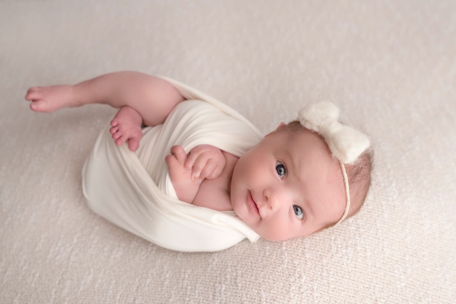A baby wrapped in white fabric lies on a soft white blanket while wearing a delicate bow headband.