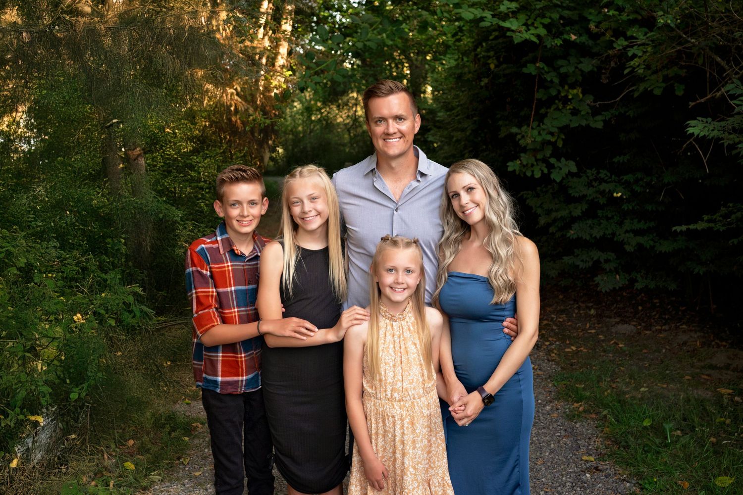 A group of five people pose together outdoors on a wooded path during golden hour lighting.
