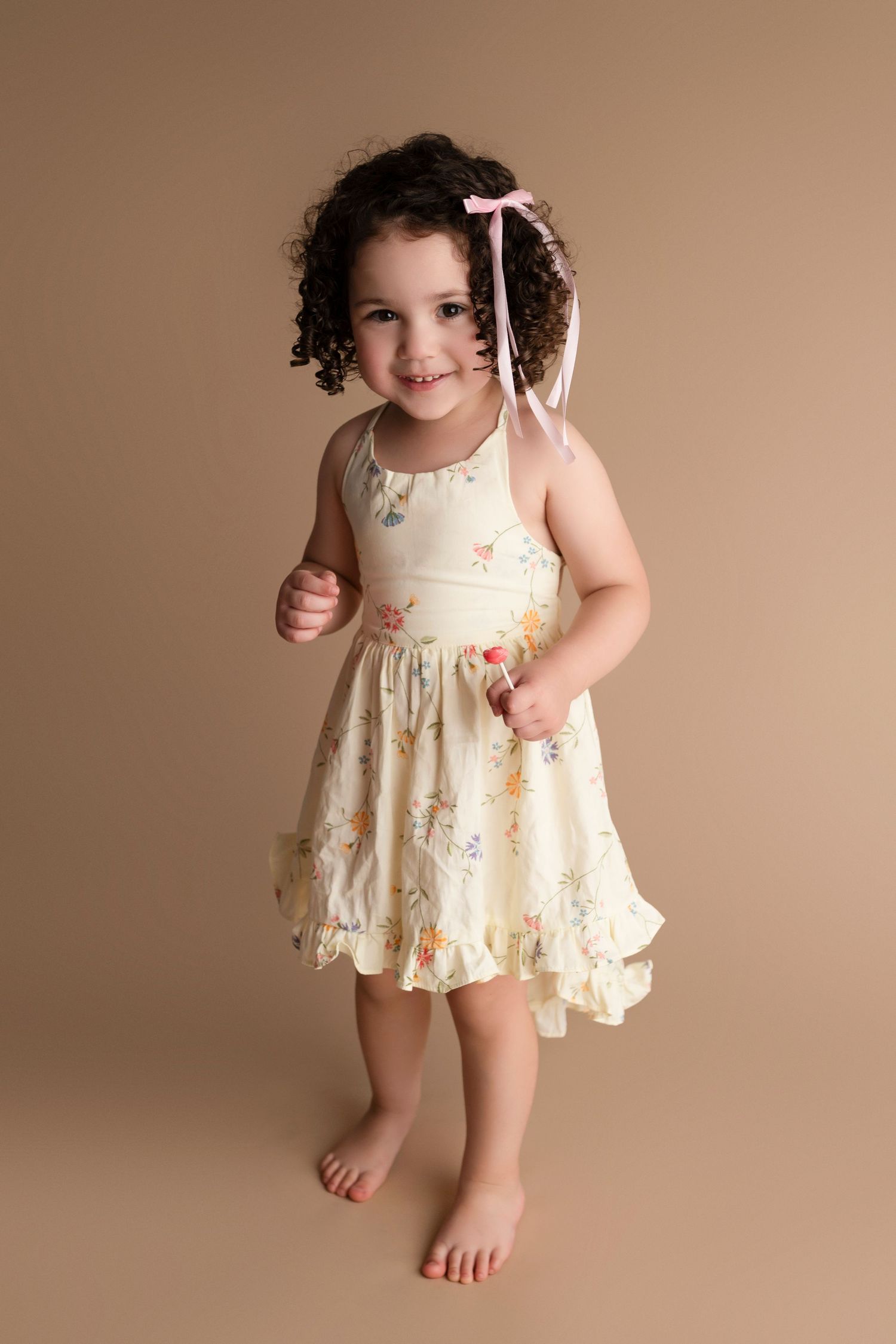Smiling toddler girl in floral dress with pink hair ribbon poses barefoot against tan studio background.