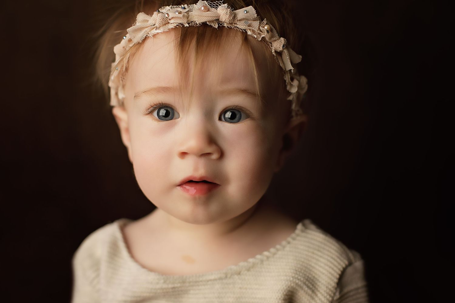 A small child wearing a delicate lace headband and cream colored sweater poses against a dark background.