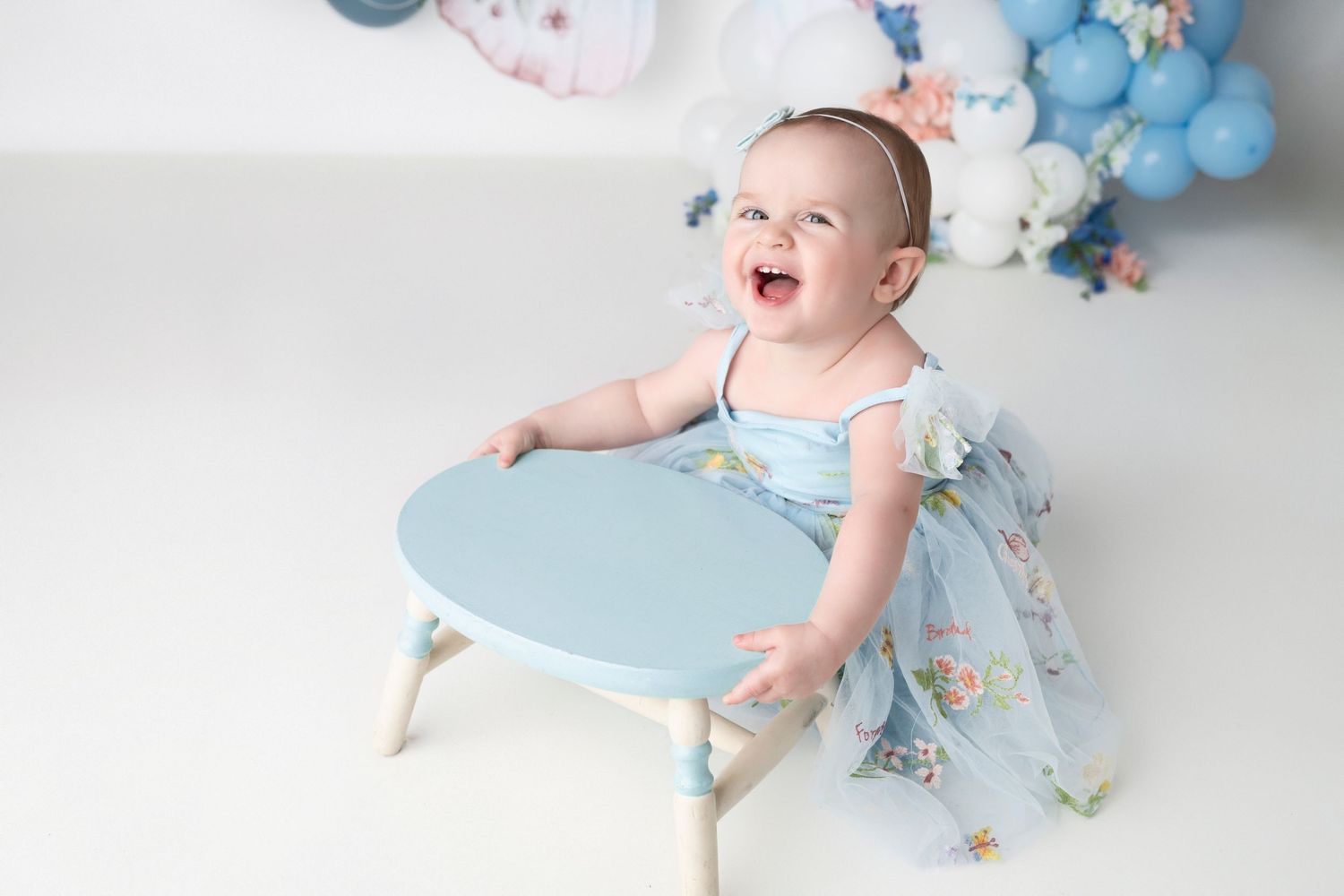 Baby in light blue outfit crawls on white backdrop with pastel balloon garland and butterfly decorations.