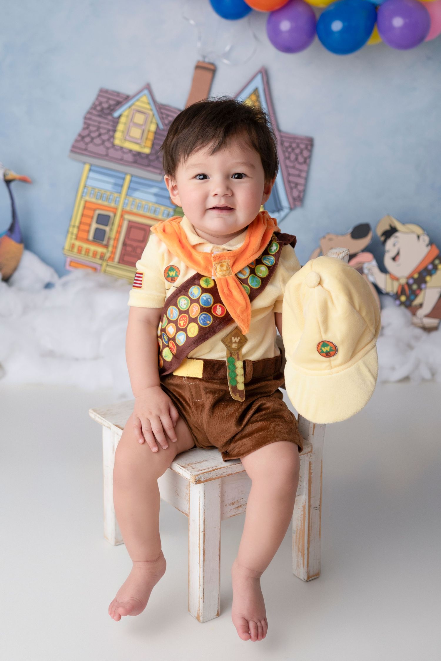 Small child in a scout uniform with badges sits on white chair against backdrop with balloons and cartoon house.