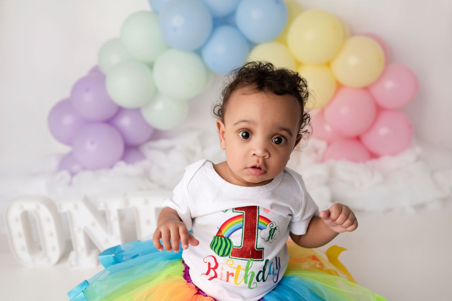 A colorful birthday celebration setup with pastel balloon garlands and rainbow tutu decorations against a white backdrop.
