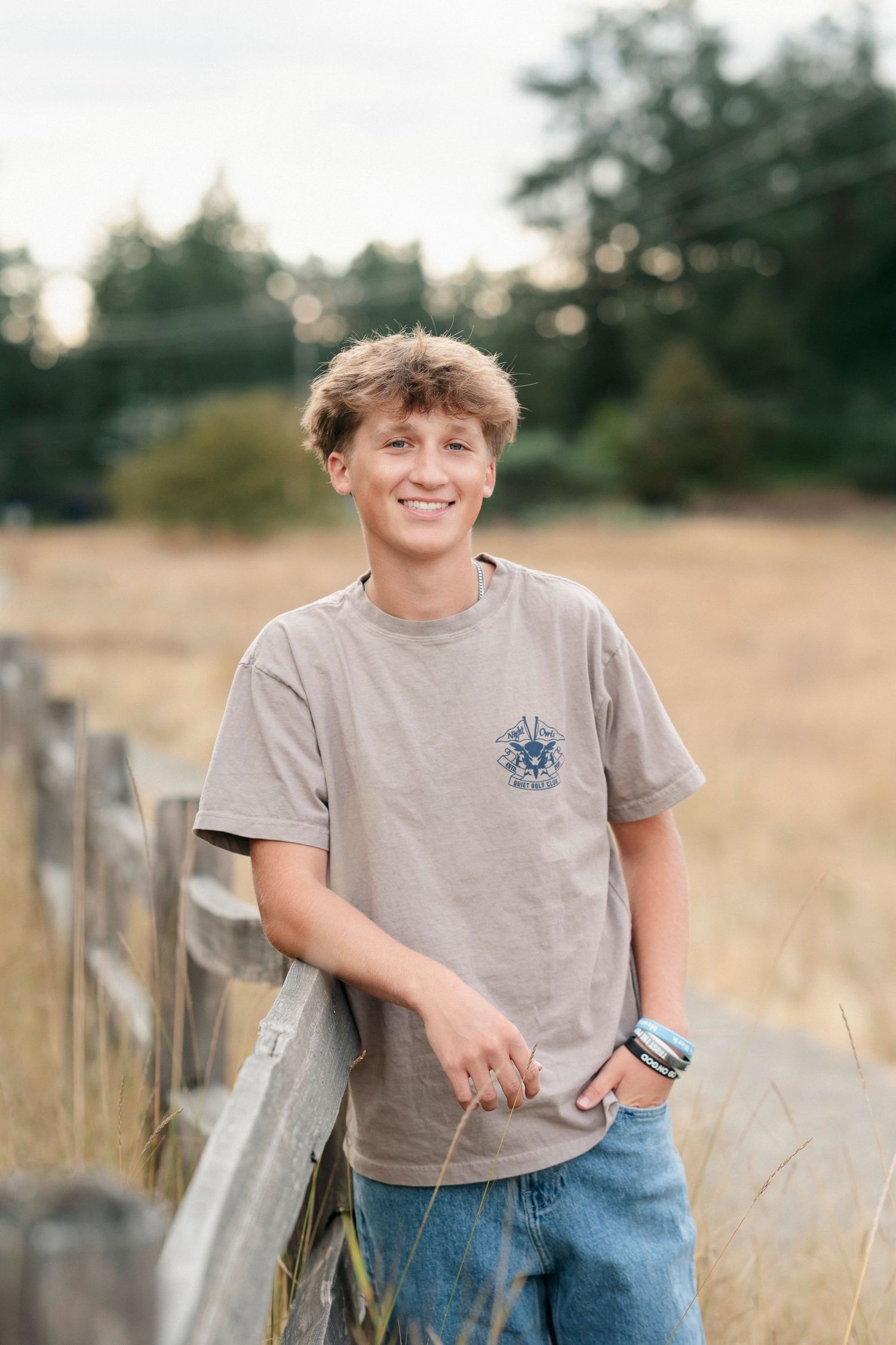 Casual portrait of someone in a gray t-shirt leaning against a wooden fence in a rural field at sunset.