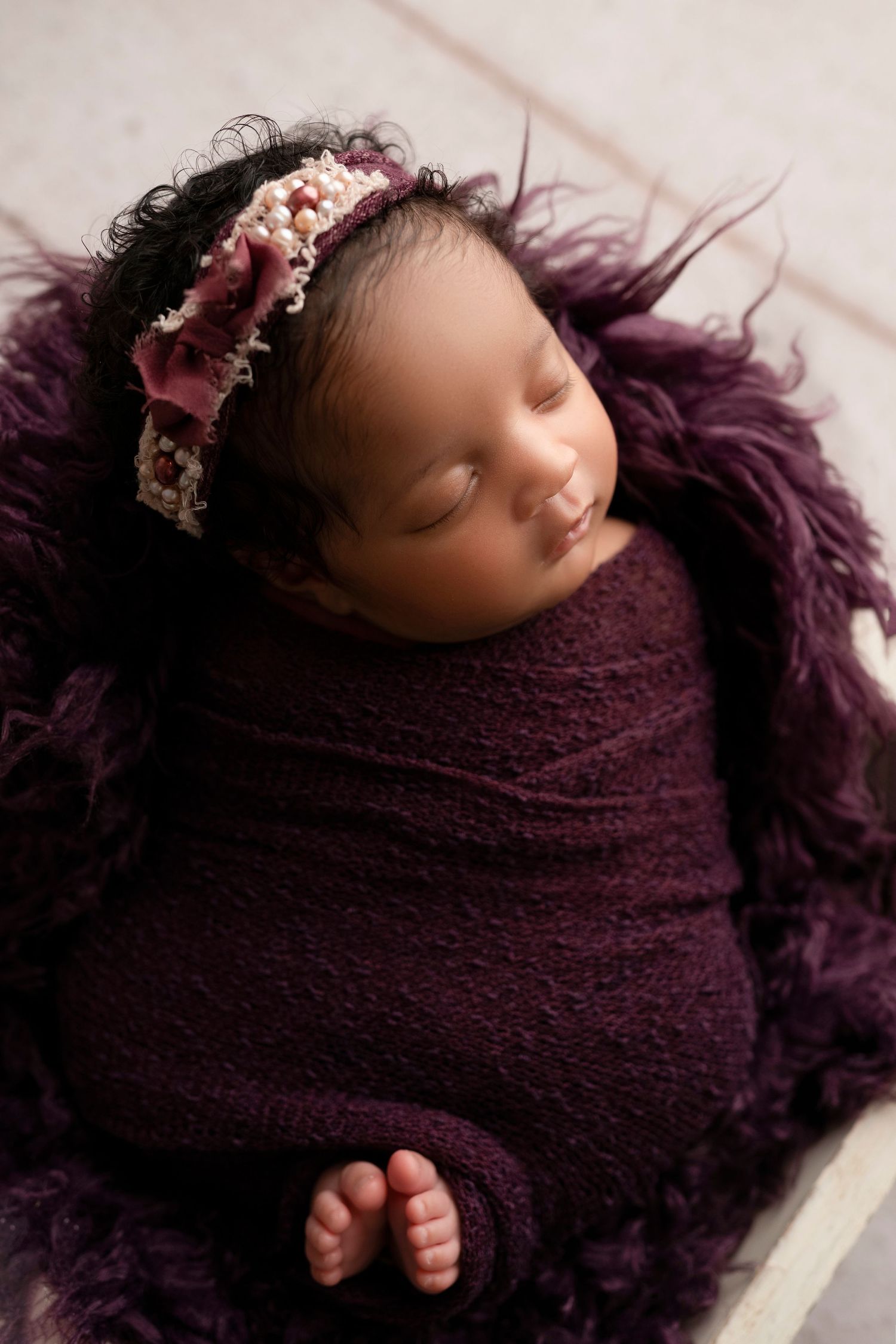 Newborn baby wrapped in purple blanket with delicate floral headpiece resting peacefully on soft textured backdrop.