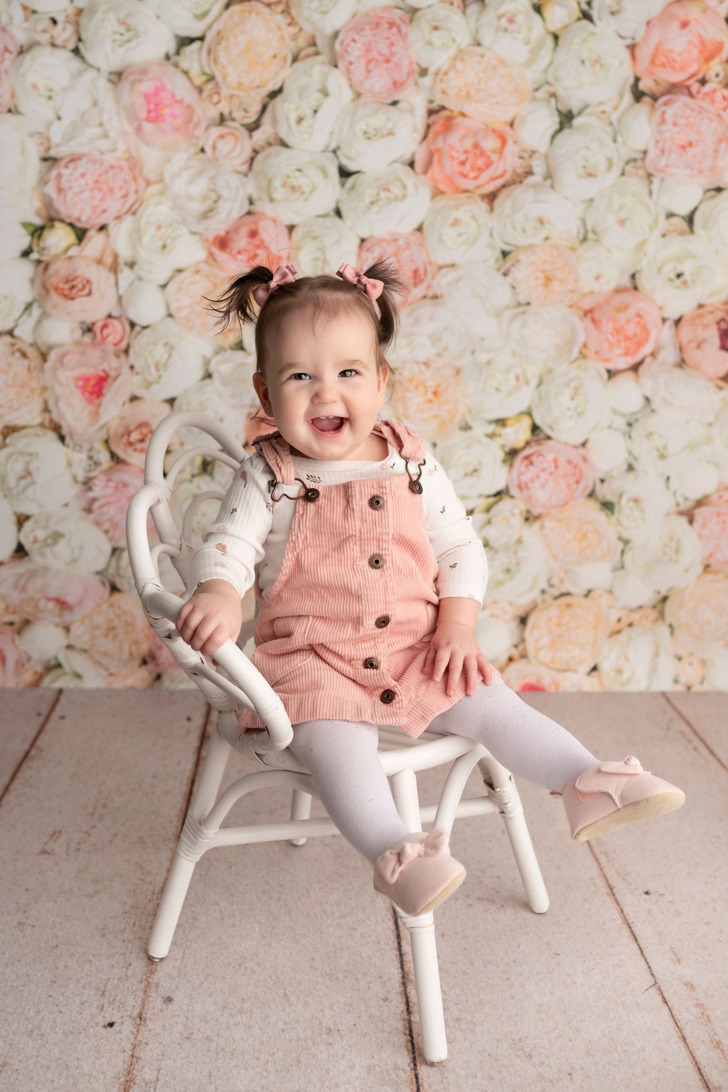 Laughing baby girl in pink dress sits on white chair against floral backdrop.