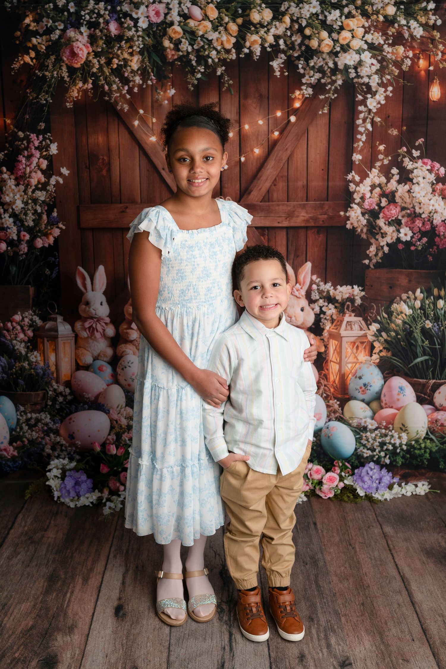 Two children pose for an Easter photo with colorful eggs, bunnies, and floral decorations against a rustic barn backdrop.
