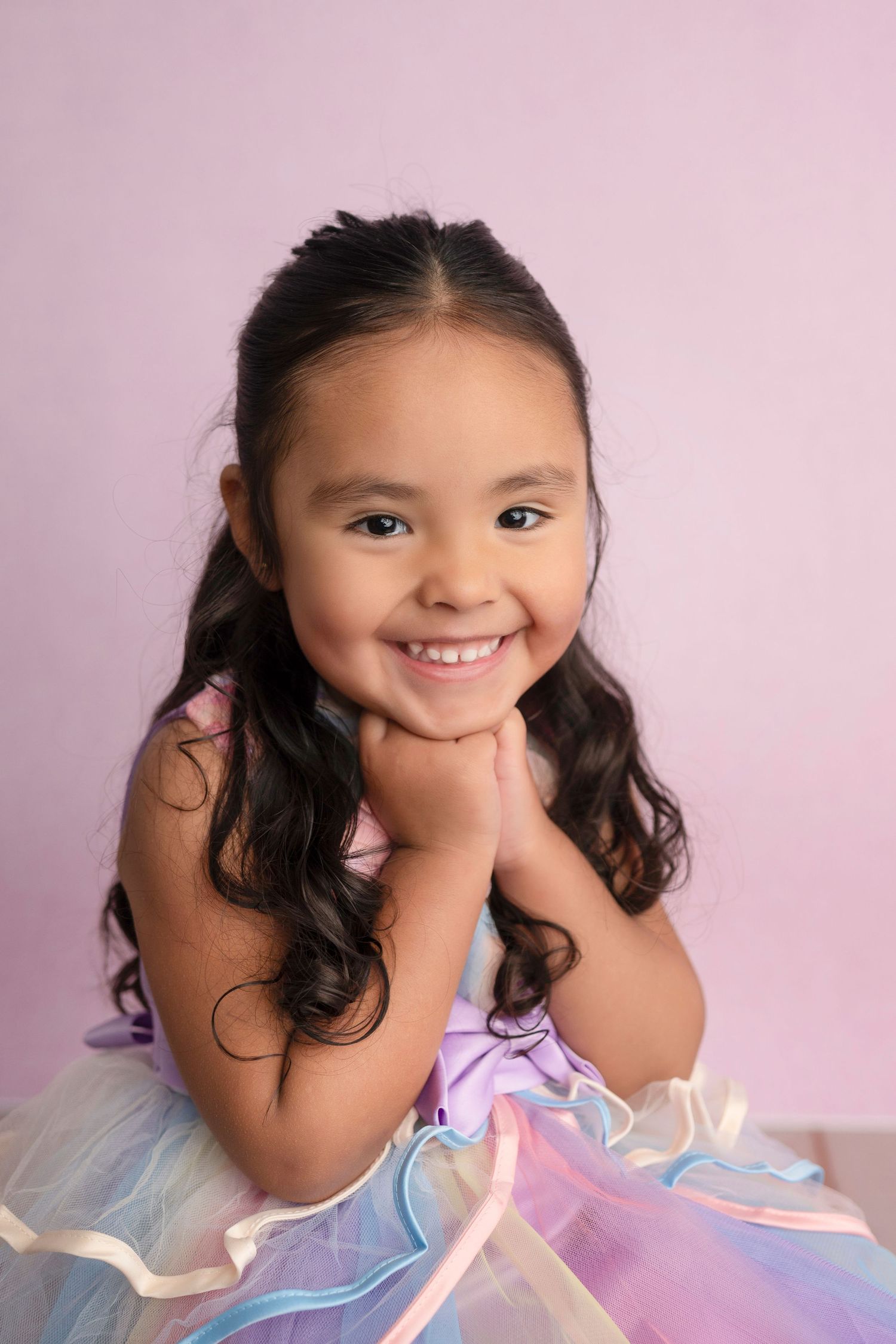 A child in a colorful tulle dress poses playfully against a pink backdrop in a studio portrait session.