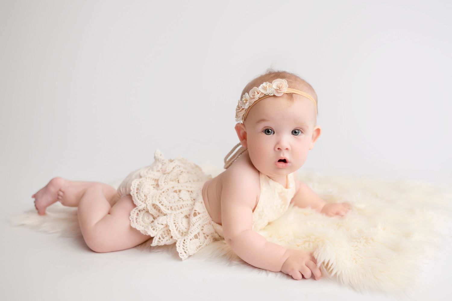 Baby in cream lace romper lying on white backdrop wearing delicate headband.